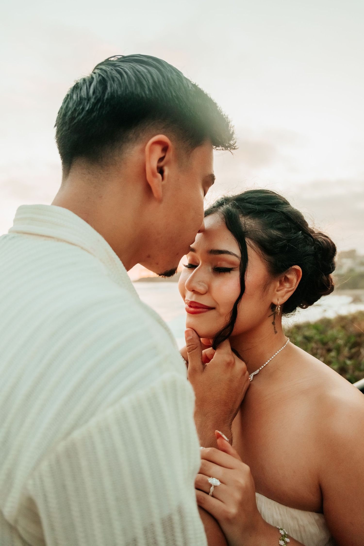 A couple shares an intimate moment outdoors, the groom tenderly cupping the bride's face as she smiles with eyes closed.