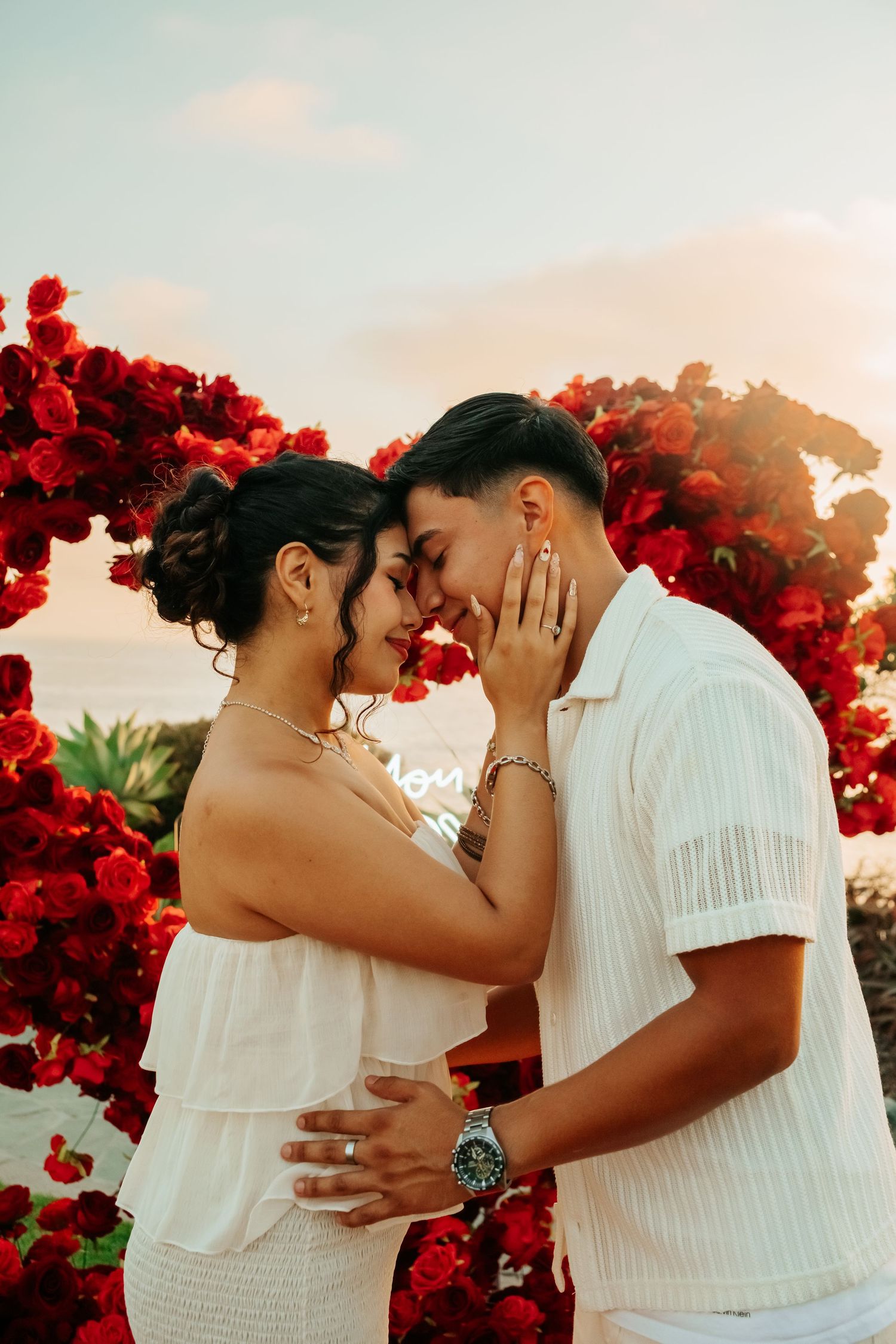 Engaged couple embracing forehead to forehead in front of red rose floral arch.