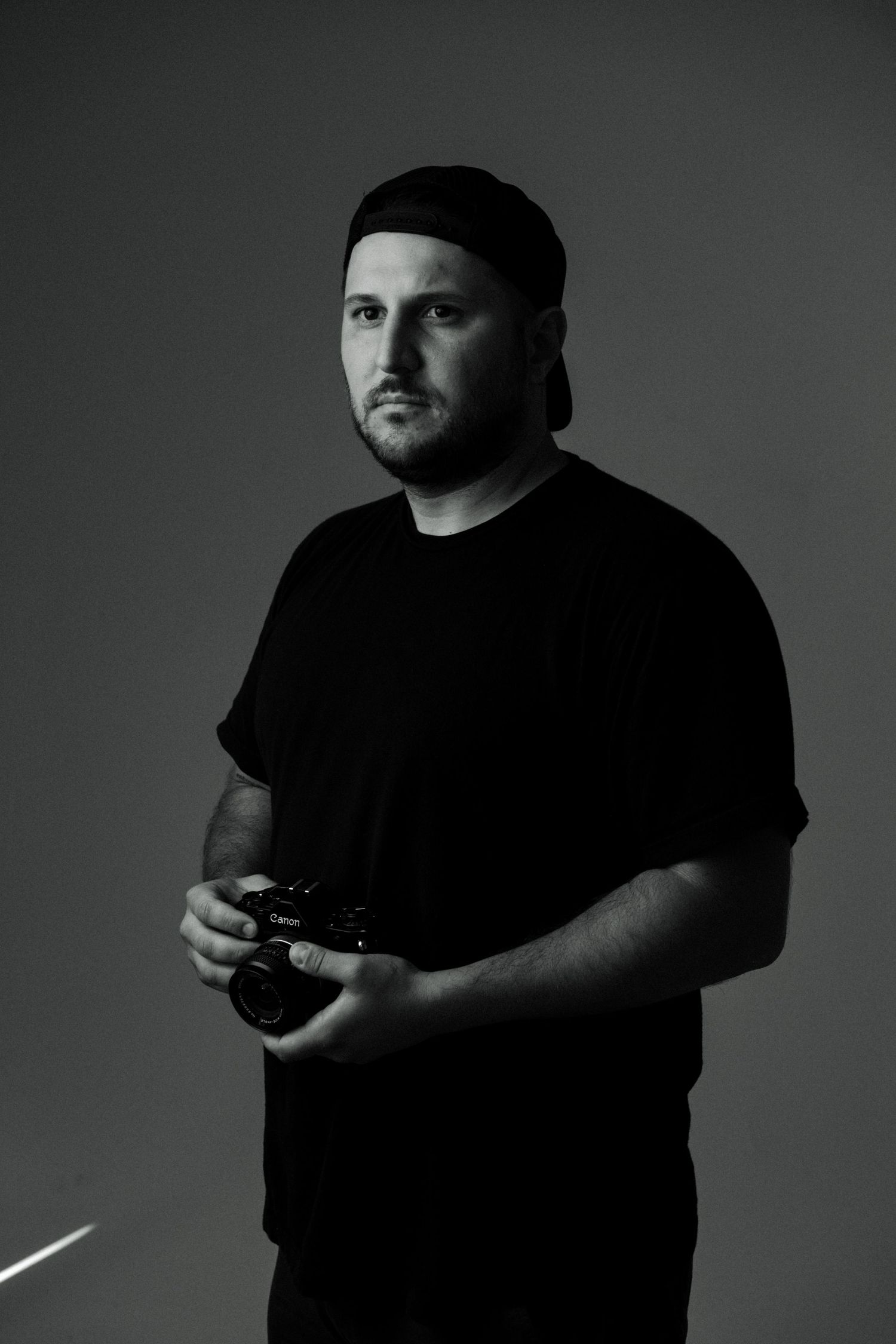 Black and white portrait of a man in a black t-shirt and cap holding a camera against a dark background.