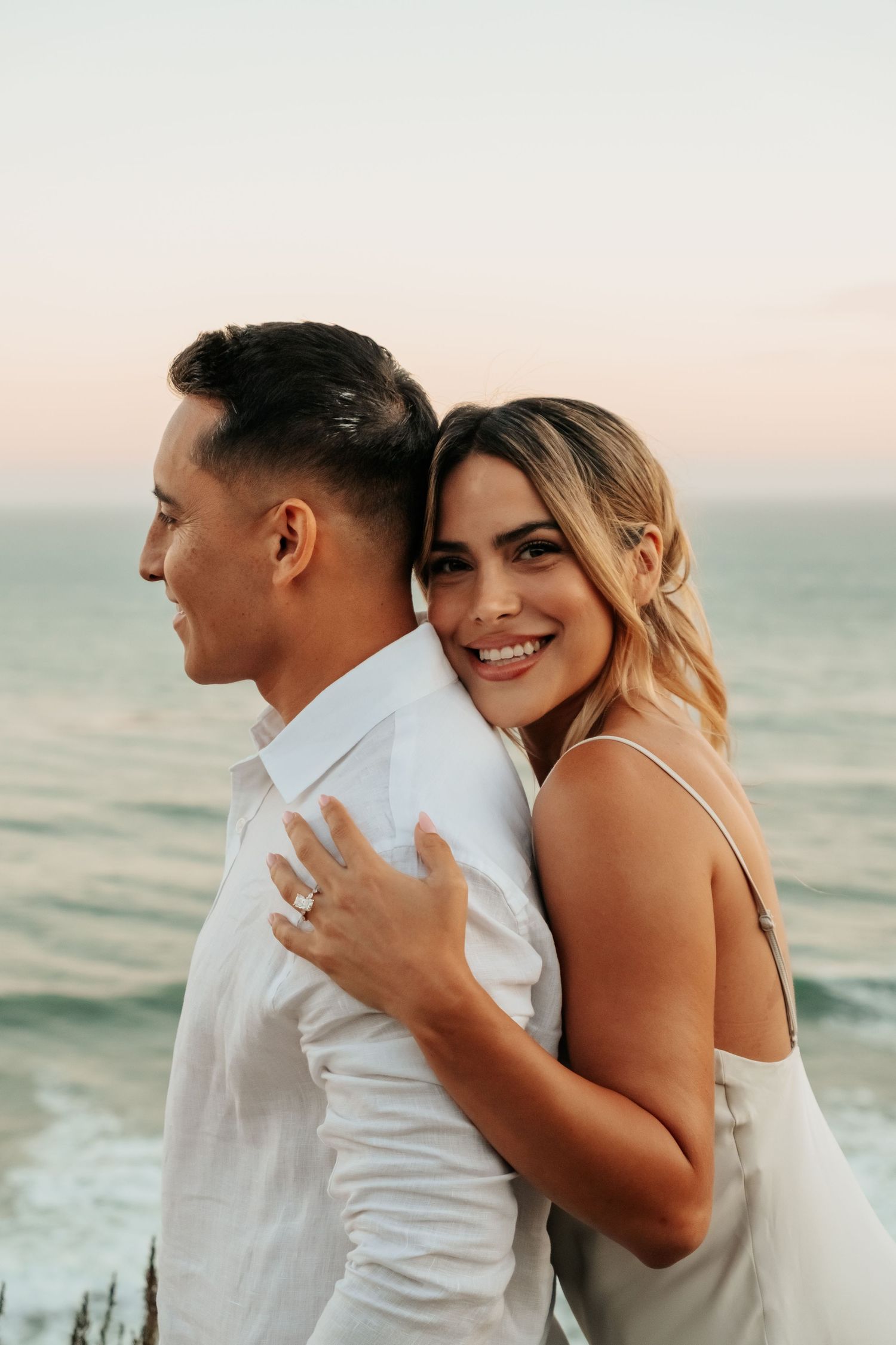 Happy engaged woman hugging man from behind smiling at camera with ocean sunset backdrop.