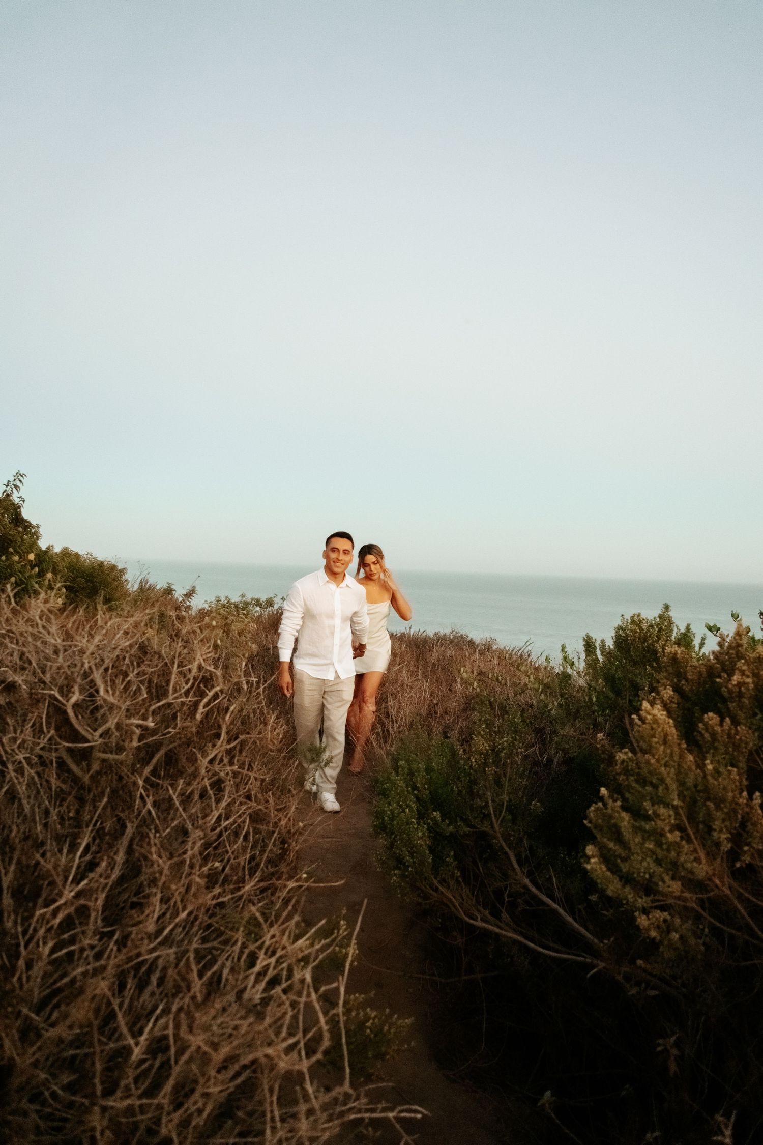 Couple in white outfits walking along narrow coastal path surrounded by shrubs.