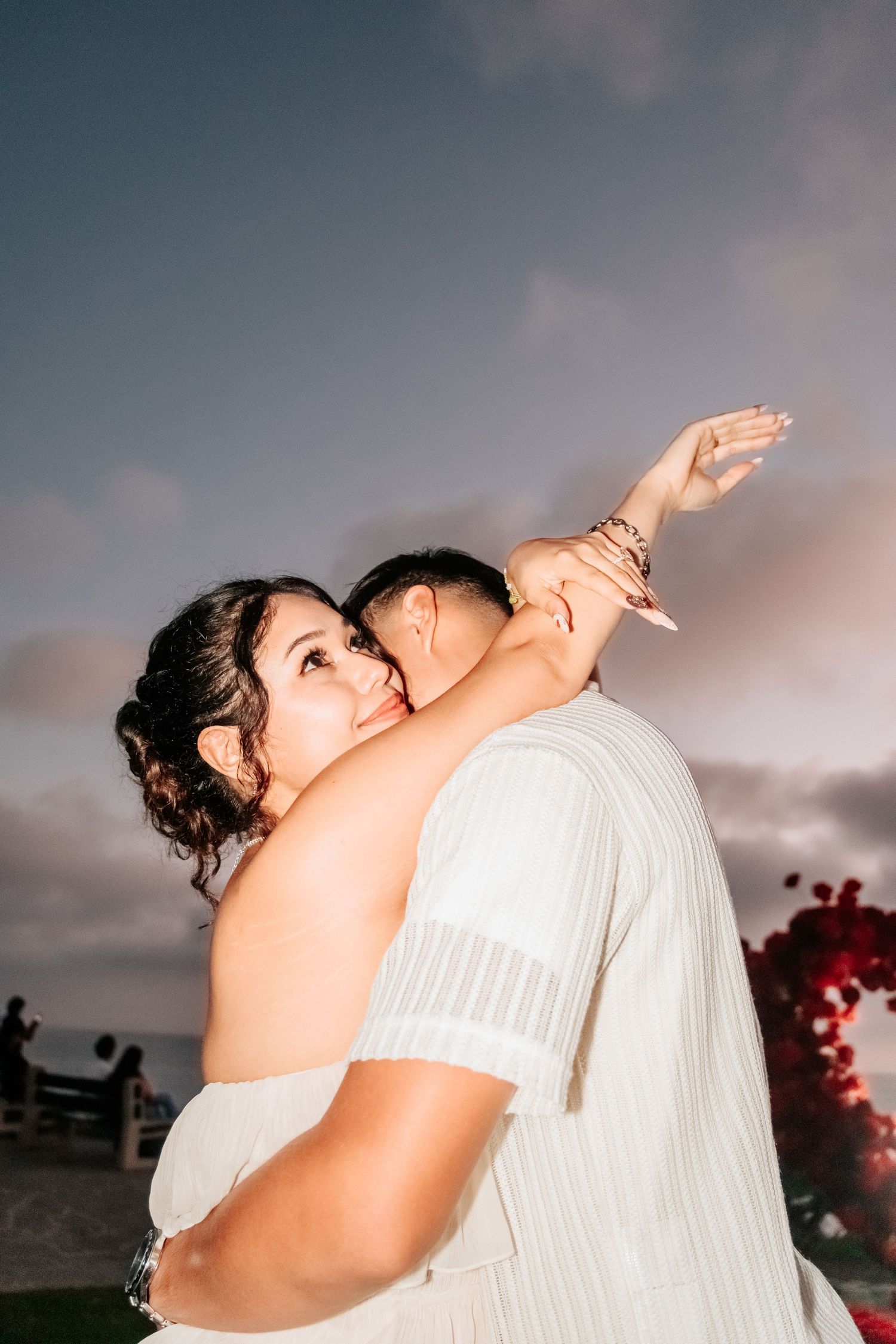 Couple embracing at sunset beach wedding, bride in white dress smiling over groom's shoulder with red florals.