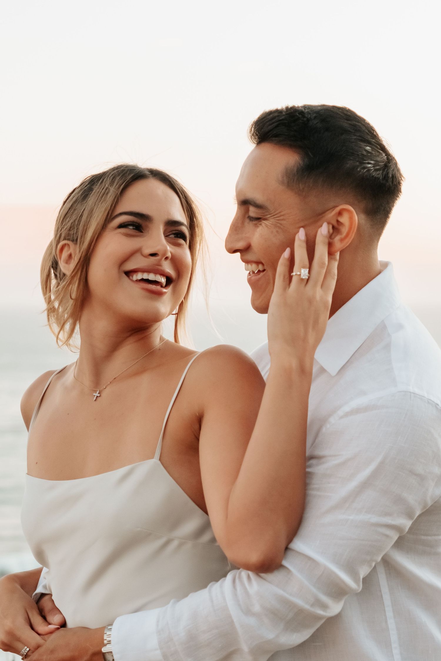 Joyful engaged couple laughing together at golden hour with sparkling diamond ring on woman's hand.