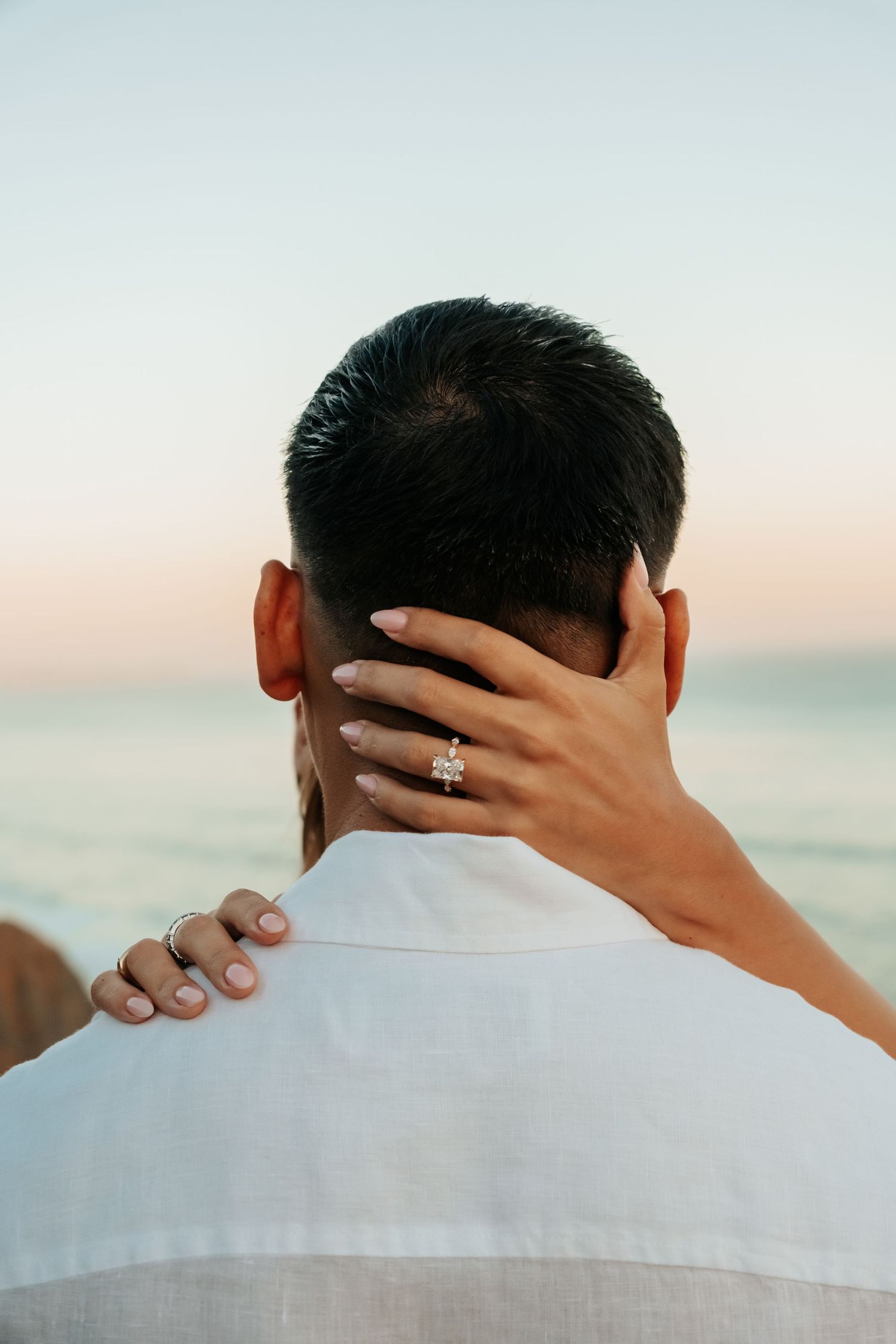 Close-up of woman's hand with stunning engagement ring resting on man's neck at sunset beach.
