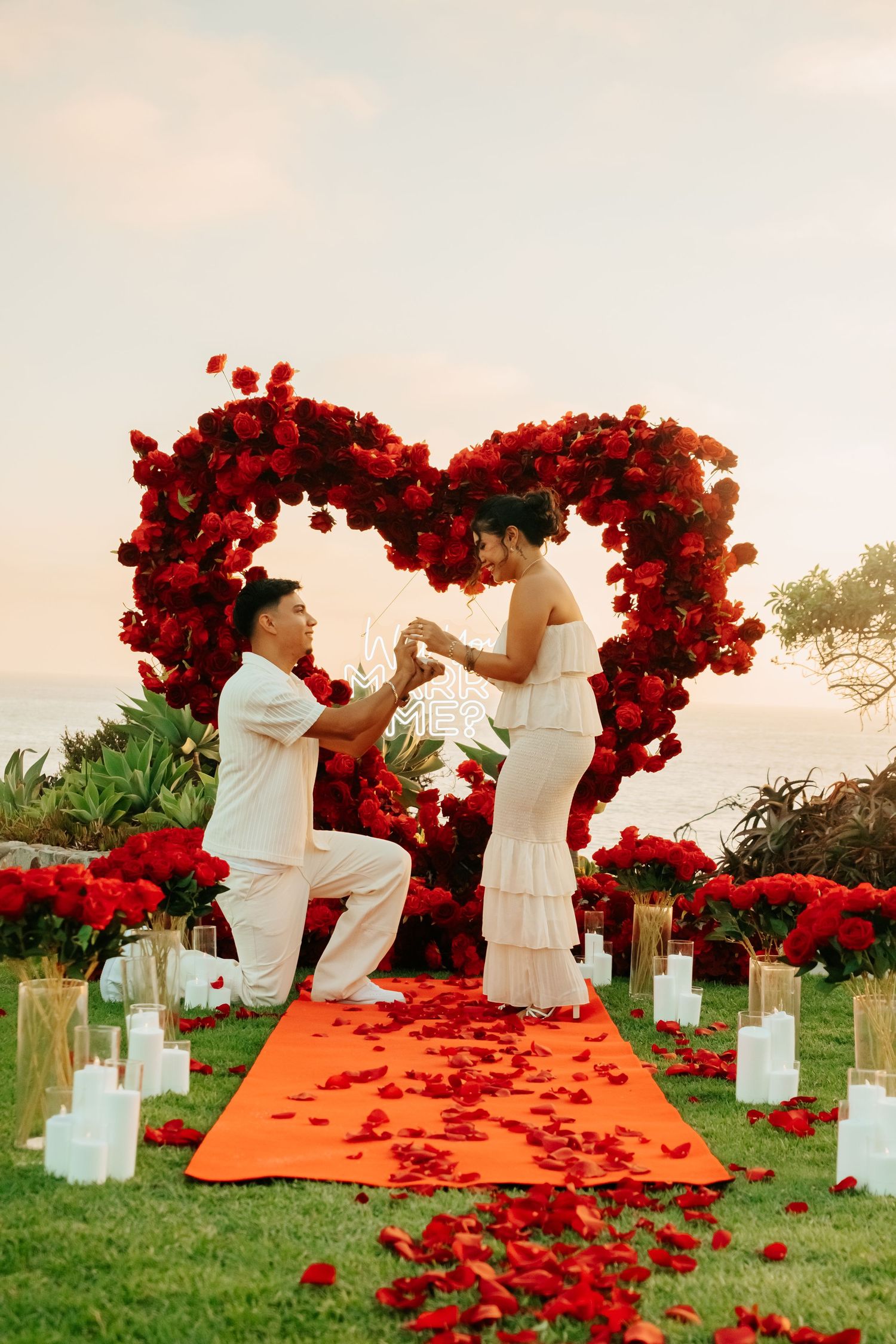 Man proposing on one knee under red rose heart arch with candles at sunset.