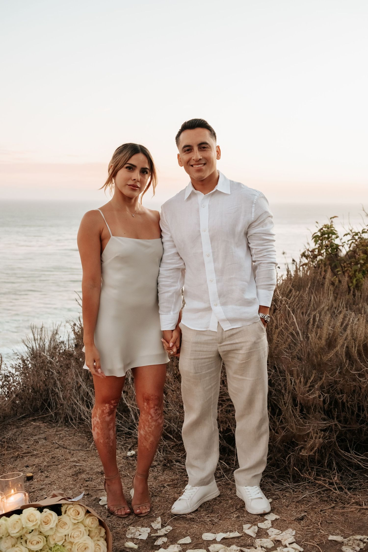 Couple in white outfits holding hands at coastal cliffside with ocean backdrop at sunset.