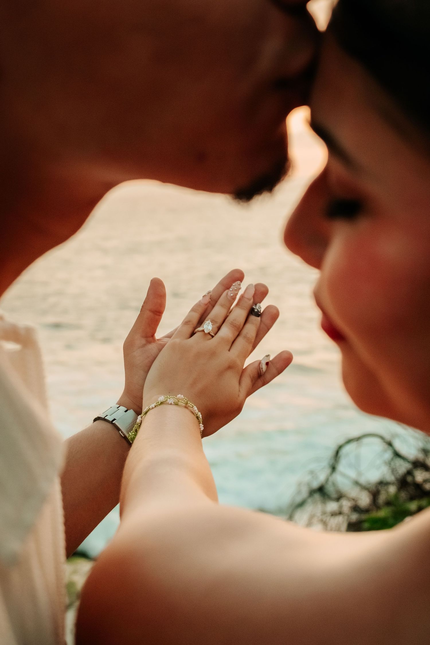 Couple sharing a romantic moment, displaying engagement ring and bracelet with ocean backdrop.