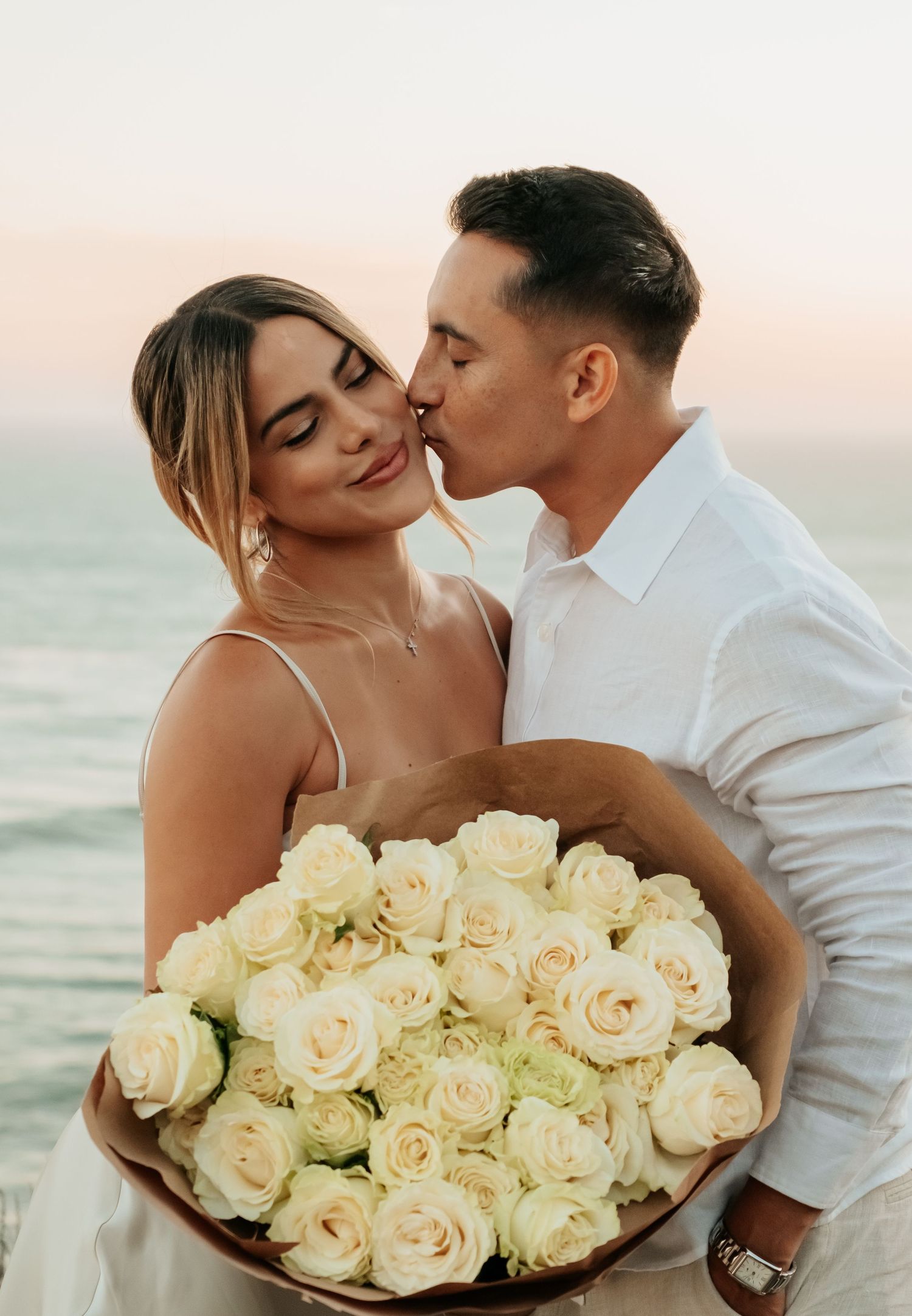 Romantic couple sharing a kiss with large bouquet of white roses at a stunning ocean sunset.