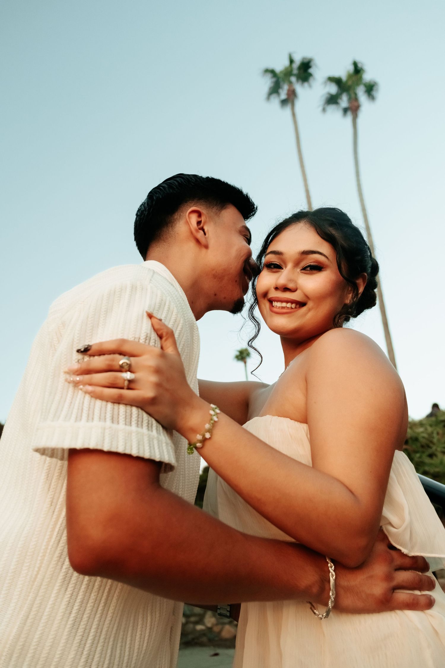 Couple embracing with man kissing woman's cheek near palm trees at dusk.