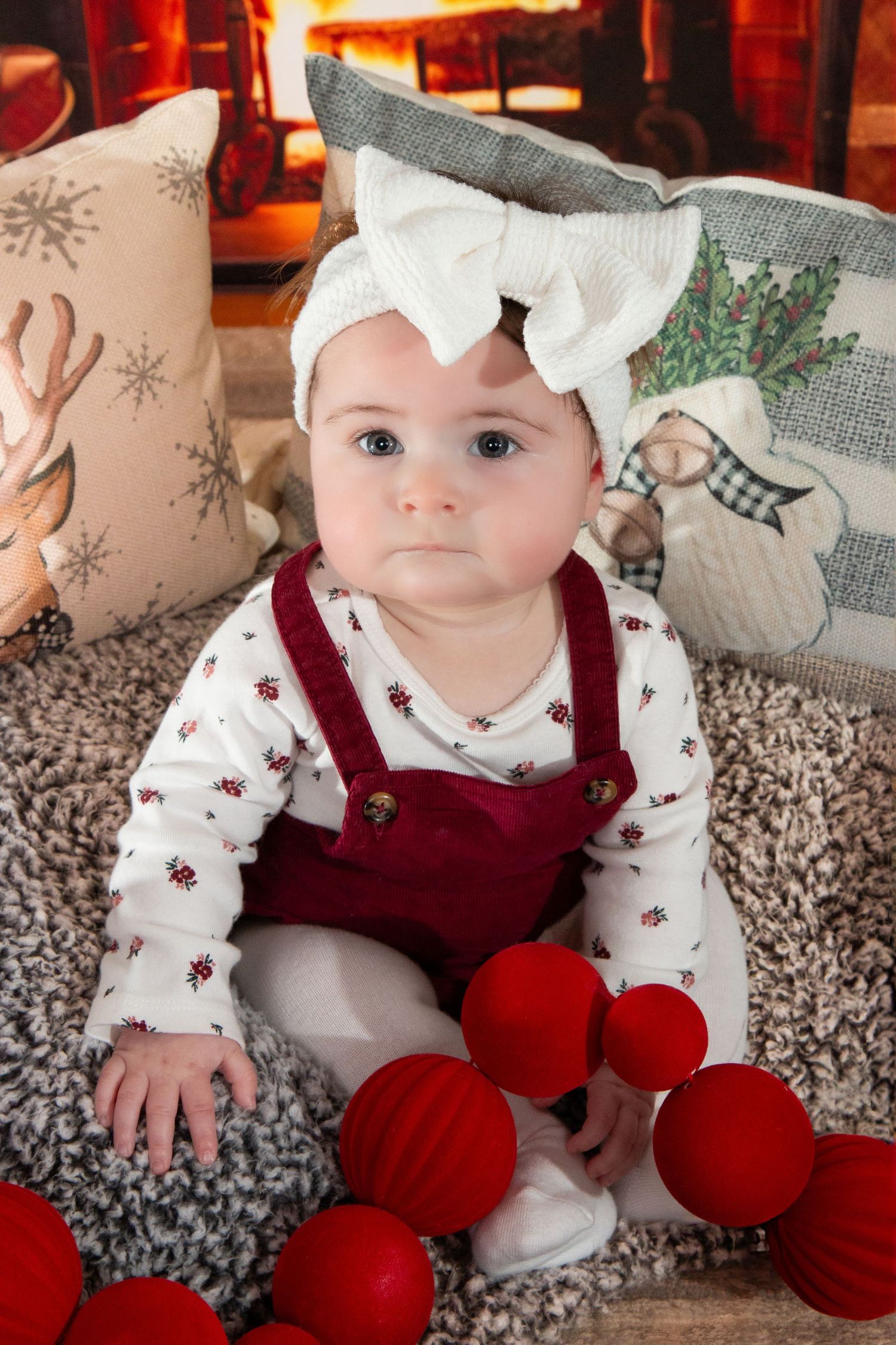 A baby girl in red velvet overalls and white bow headband sits on a rug near a warm fireplace.
