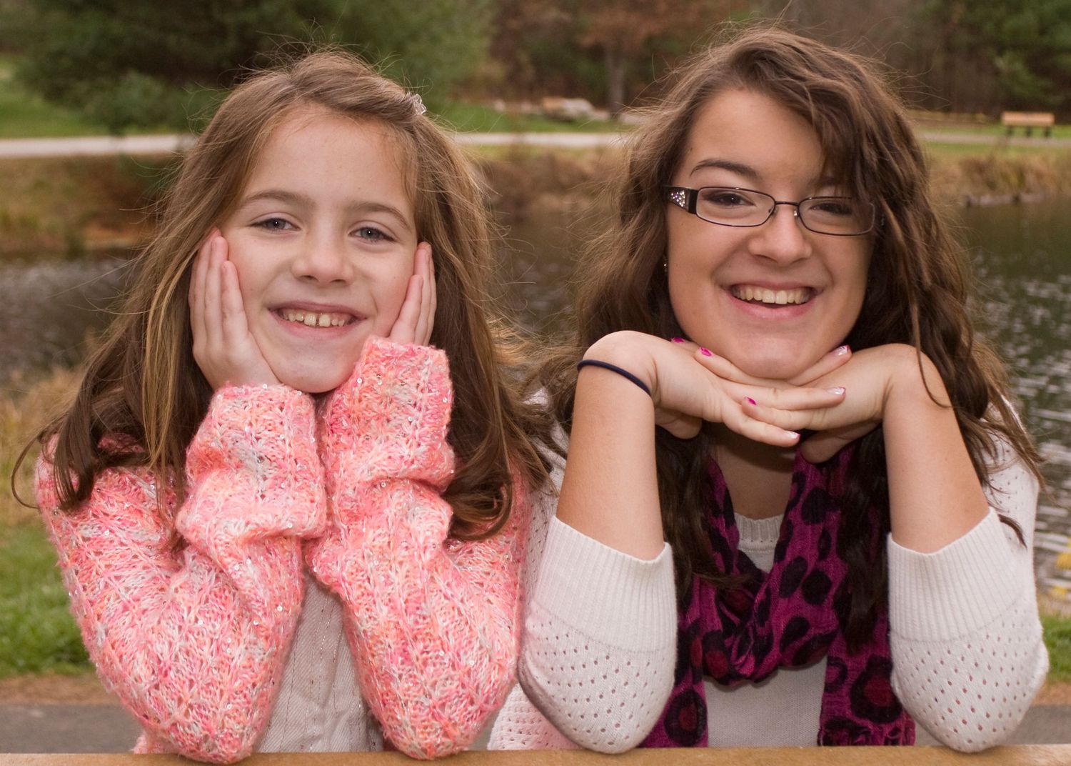 Two smiling sisters pose chin-in-hands together near a pond in outdoor portrait.