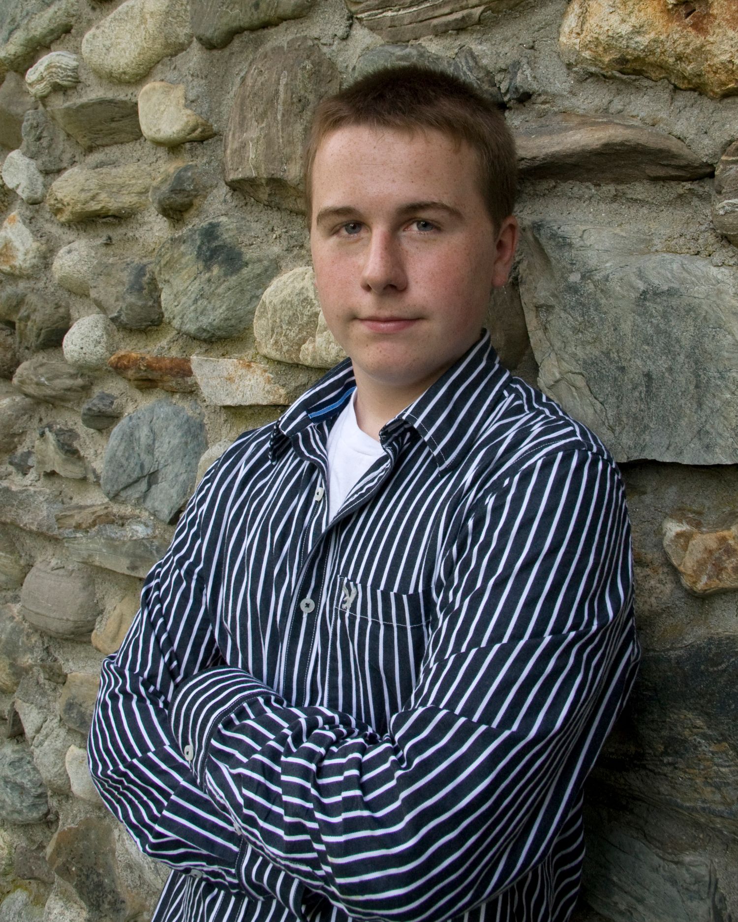 Young man in a striped shirt leaning against a rustic stone wall with arms crossed.