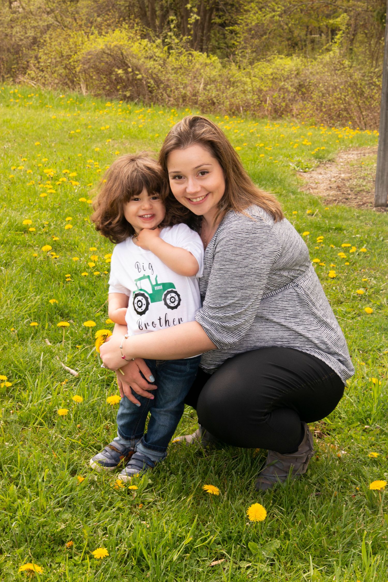 A smiling woman crouches in a dandelion field hugging a young boy wearing a 'Big Brother' tractor shirt.