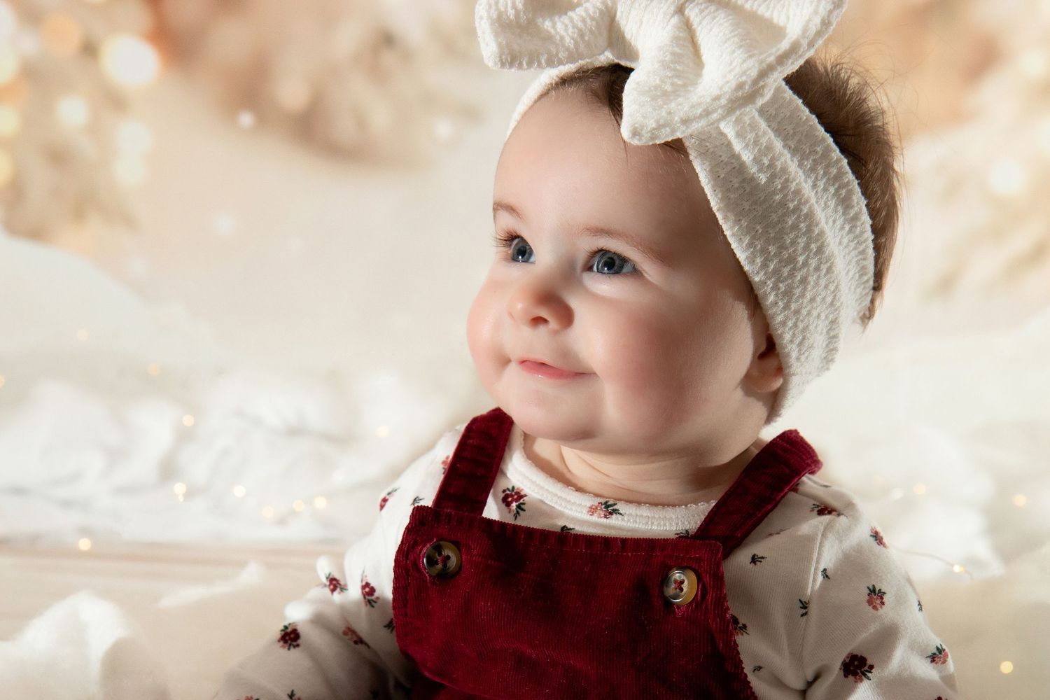 An adorable baby girl in a red velvet outfit and white bow headband gazes upward in a soft dreamy scene.
