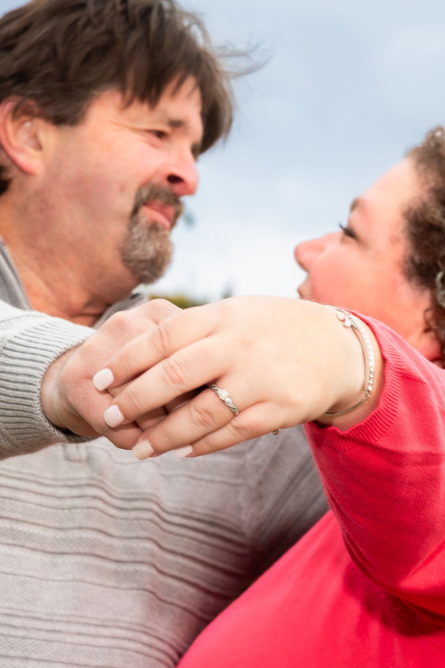 Couple holds hands showing engagement ring while gazing at each other outdoors on a cloudy day.