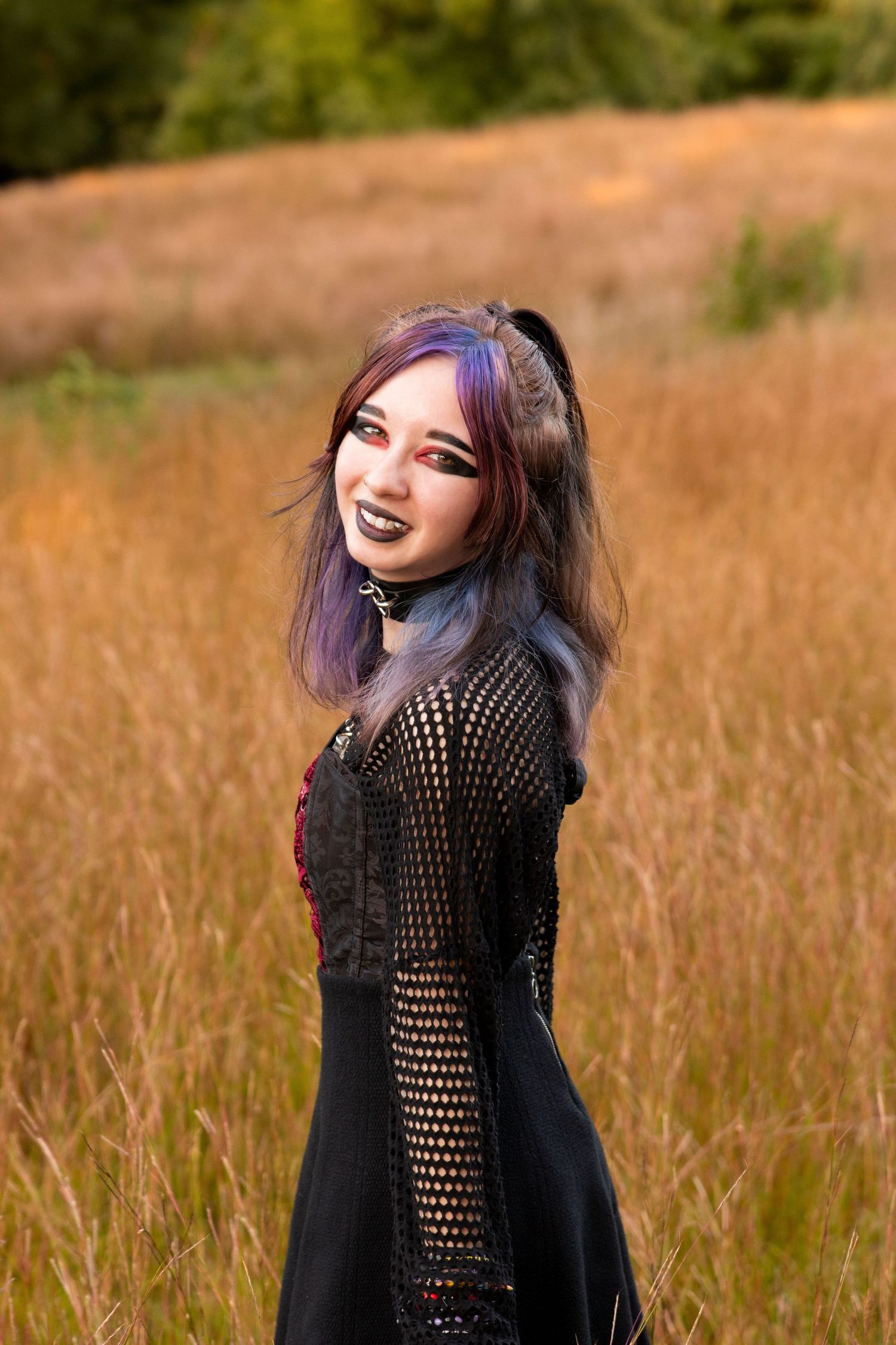Teen girl with purple hair in gothic black mesh dress standing in a golden autumn field.