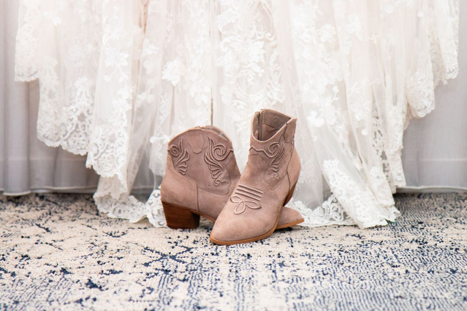 Tan cowboy boots placed in front of lace wedding dress on blue patterned rug.