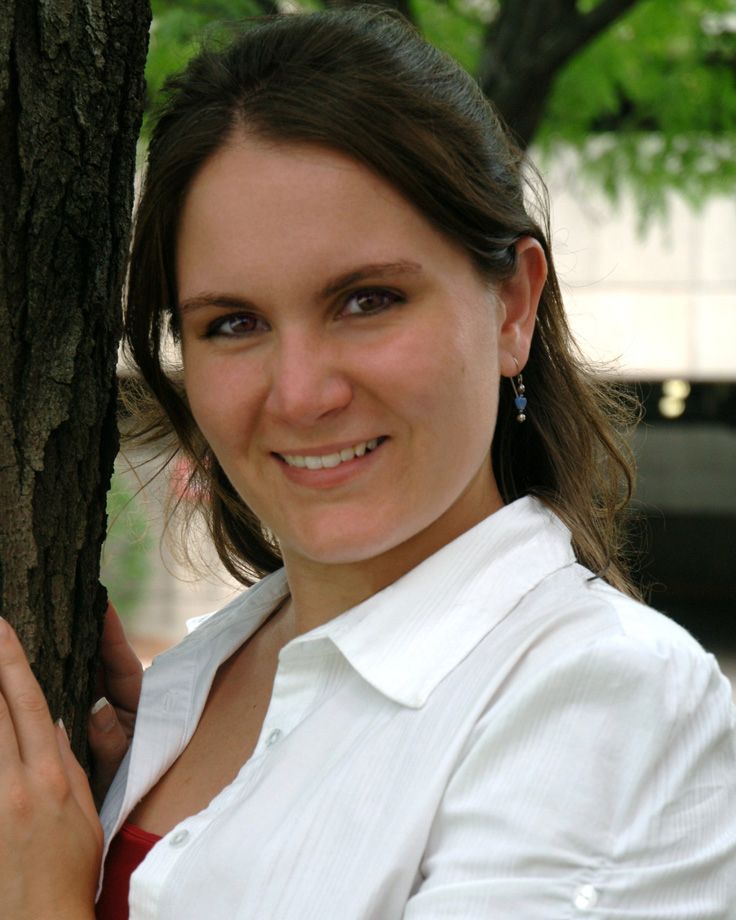 Smiling young woman with brown hair in white shirt leaning against tree outdoors.