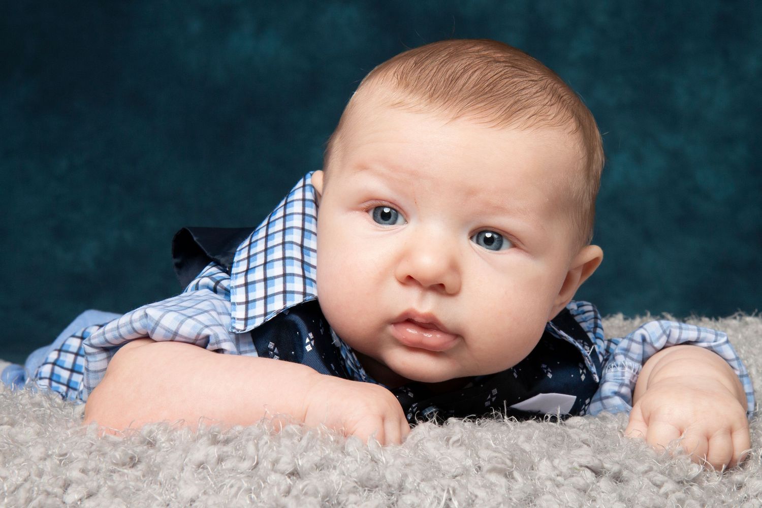 Adorable baby boy in blue plaid shirt lying on gray fluffy carpet looking at camera.