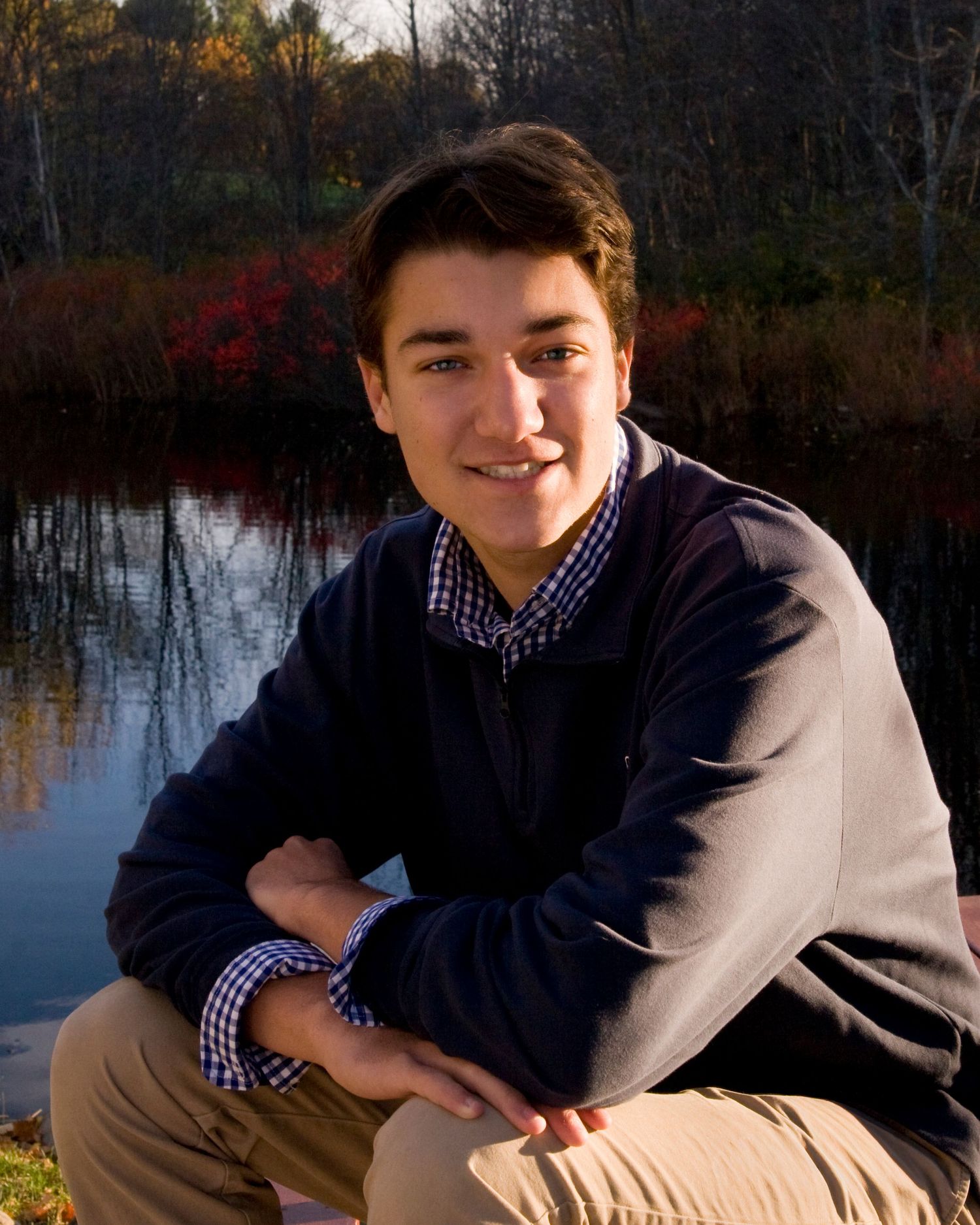 Smiling young man in a navy sweater crouching by a reflective pond surrounded by fall foliage.