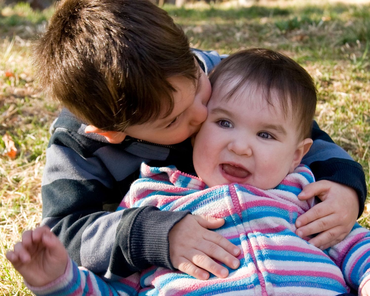 Young boy kissing baby sibling on cheek while holding them outdoors in autumn setting.