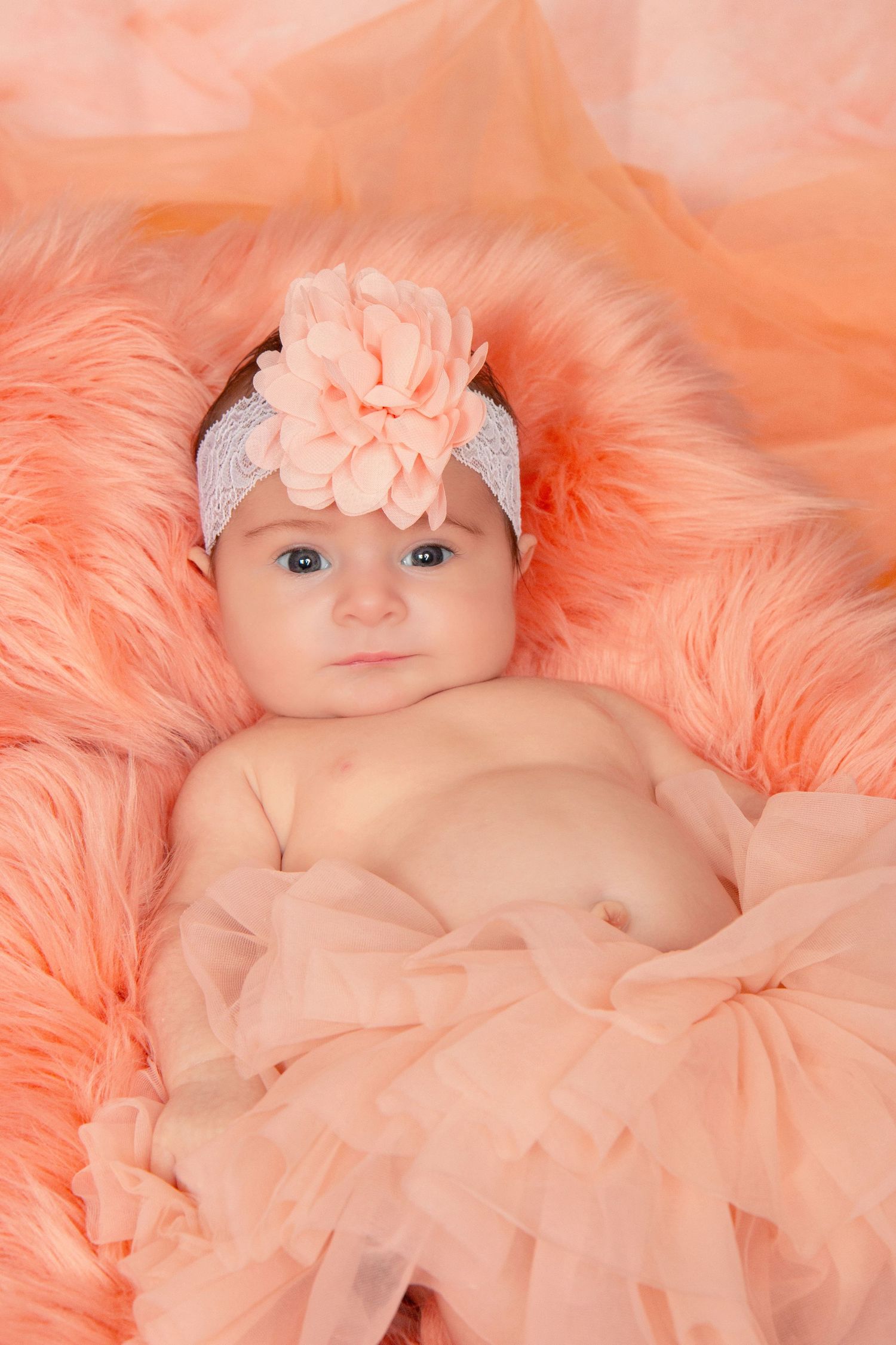 Newborn baby girl lying on pink fur with peach floral headband and tulle skirt.