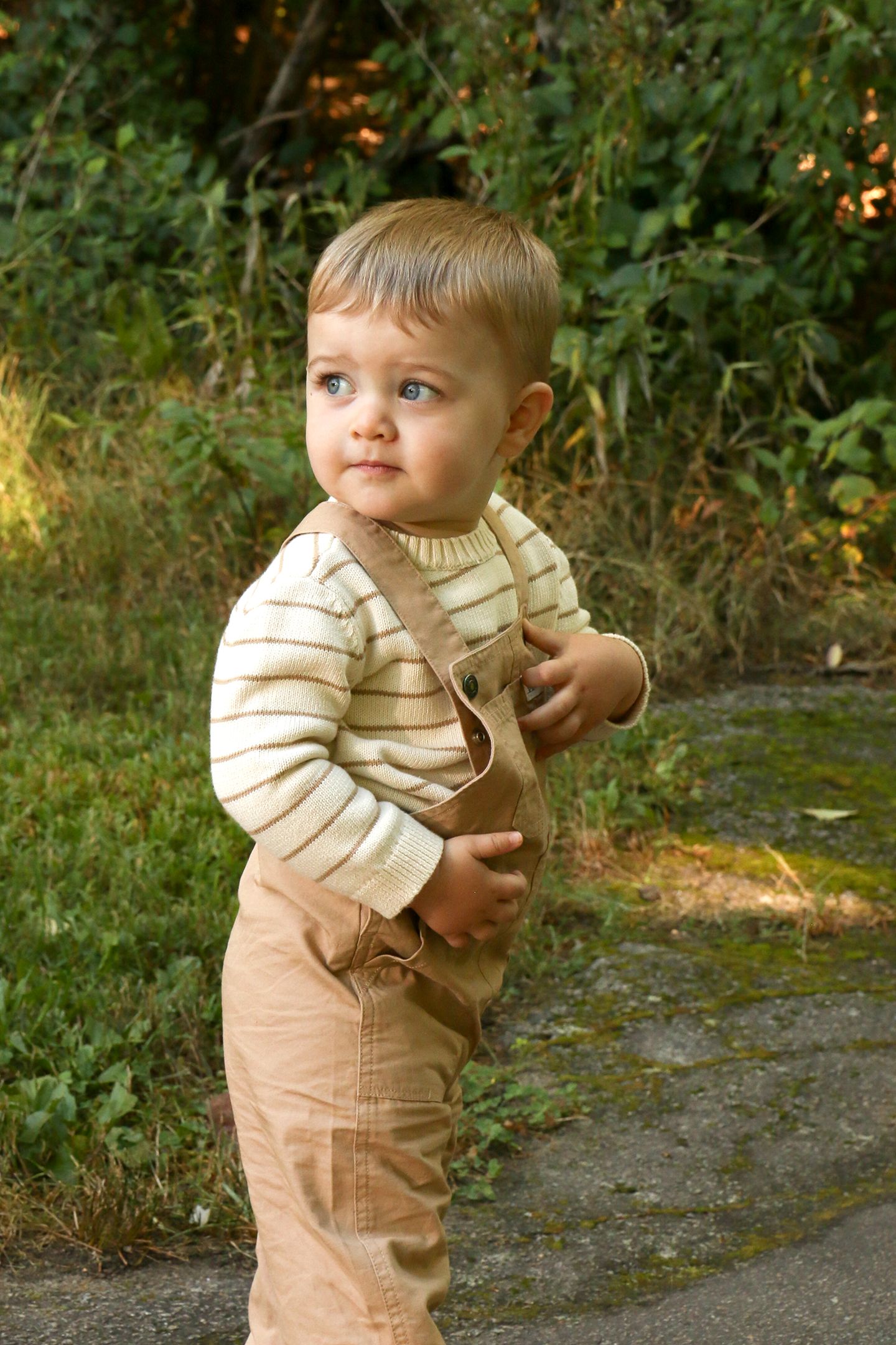 An adorable toddler in striped sweater and tan overalls stands on a garden path looking to the side.