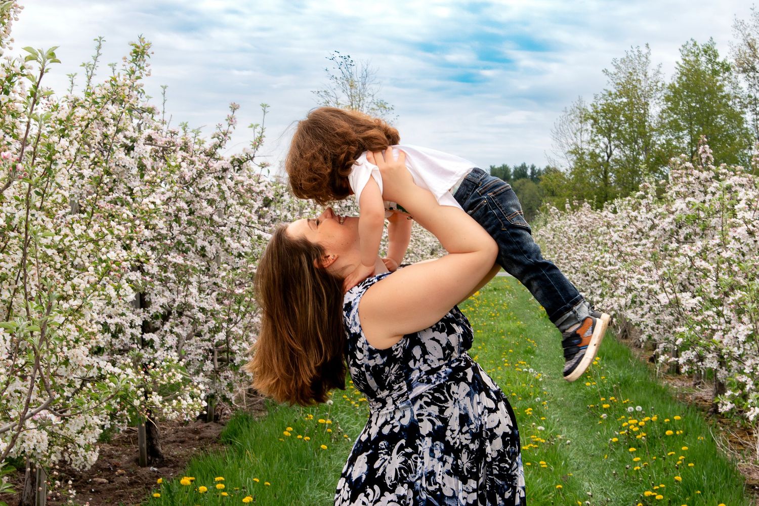 A mother joyfully lifts her young child into the air among blooming white apple trees in spring.