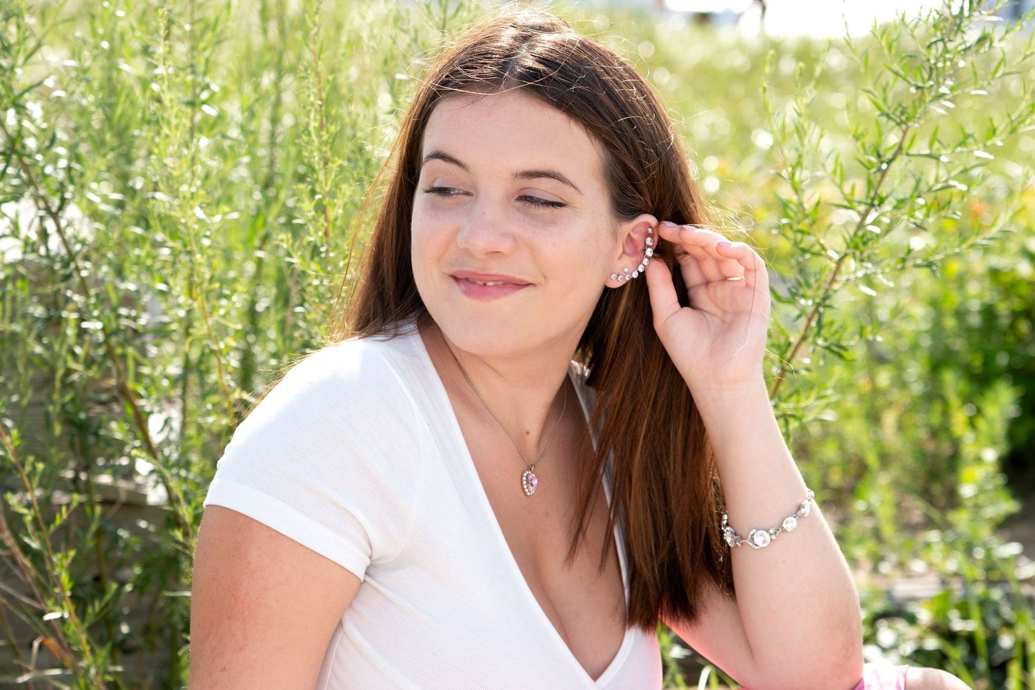 Young woman with brown hair wearing a white top and jewelry sitting among green shrubs outdoors.