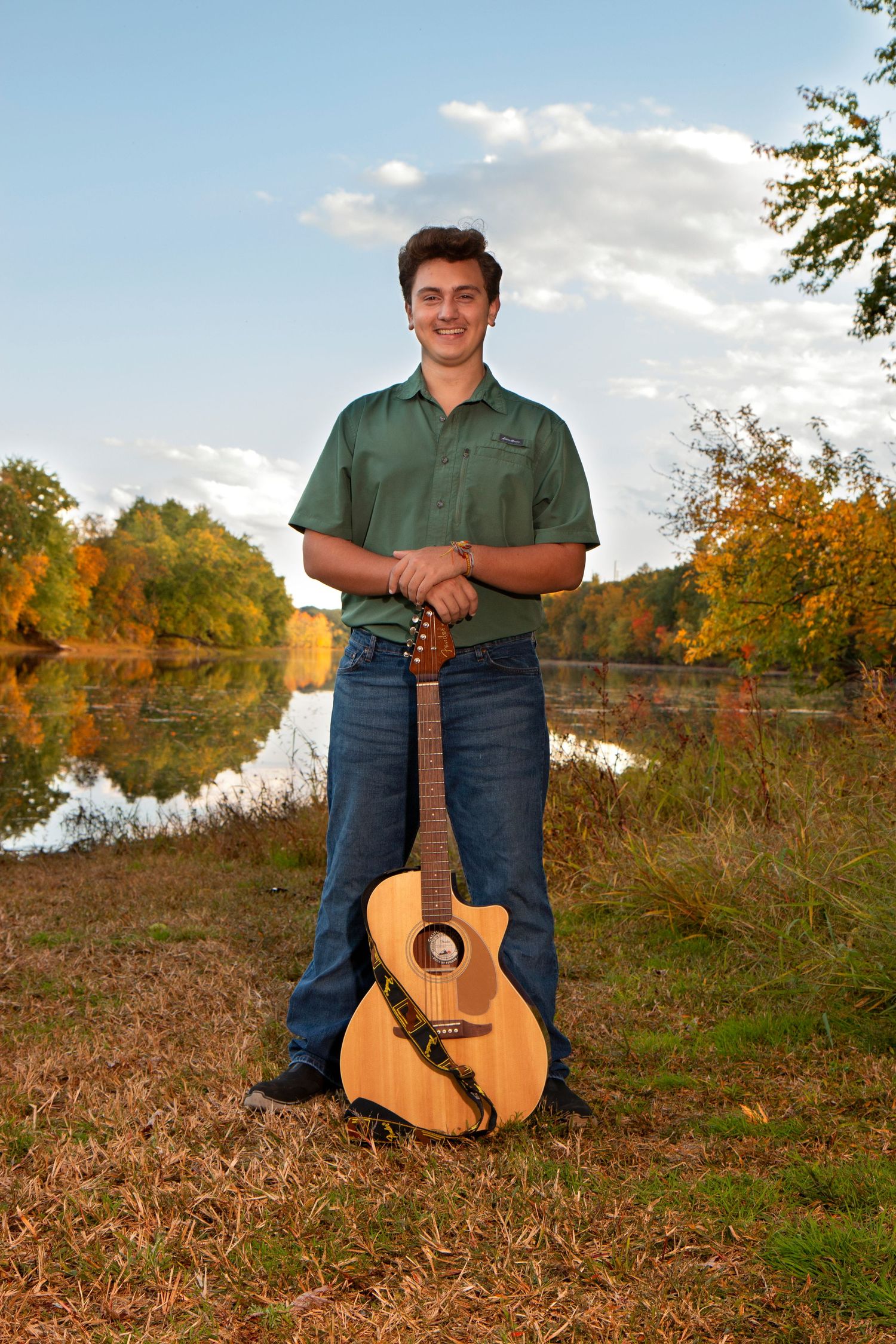 Young man in a green shirt posing with an acoustic guitar by a scenic autumn lakeside at sunset.