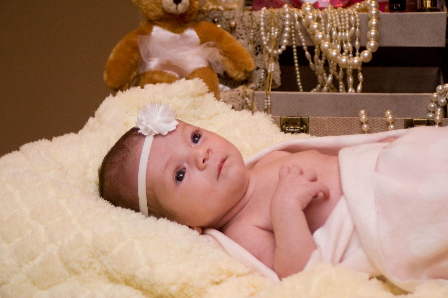 Newborn baby girl with white bow headband wrapped in pink blanket near teddy bear.