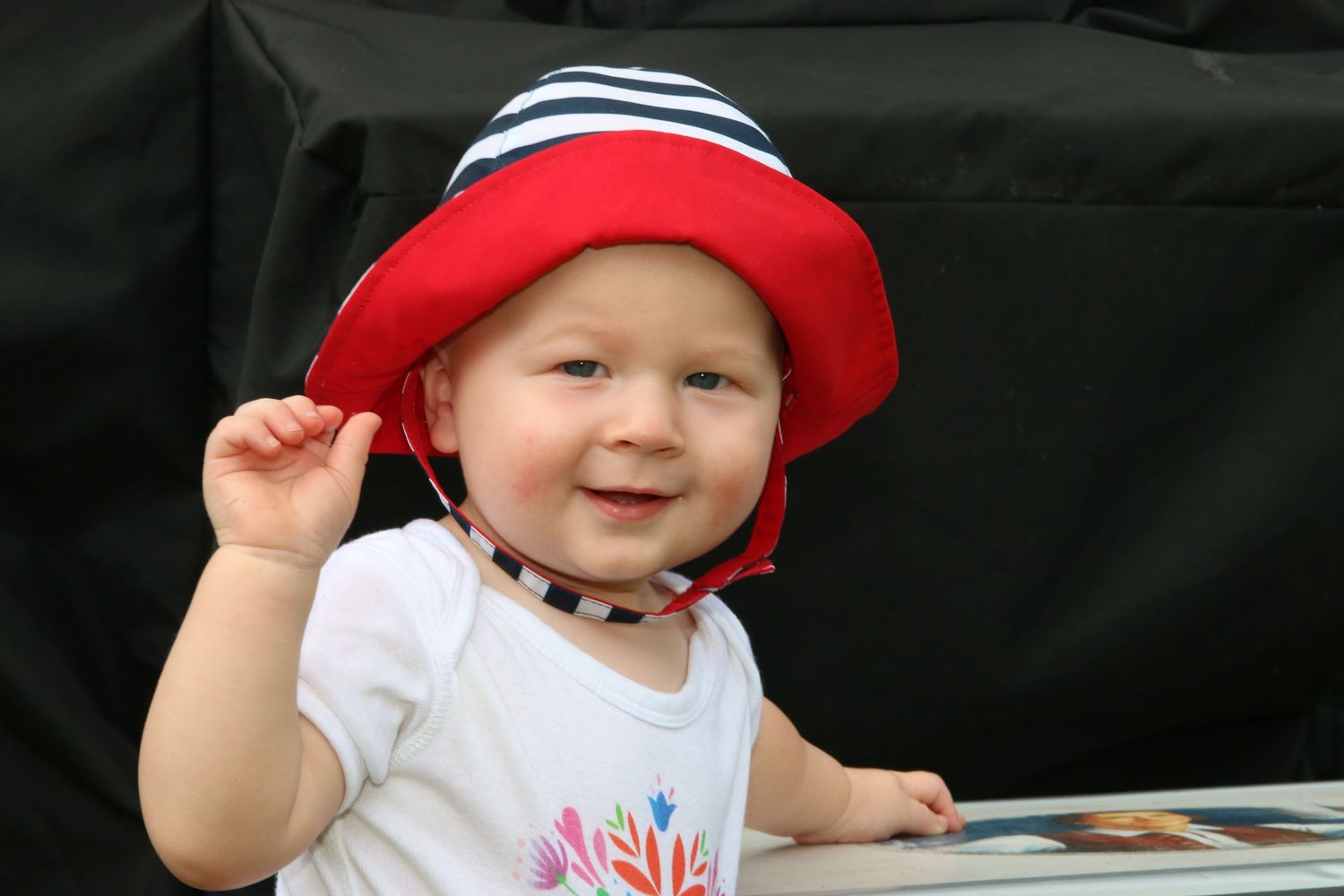 Happy baby wearing a red and navy striped bucket hat smiles brightly at the camera.
