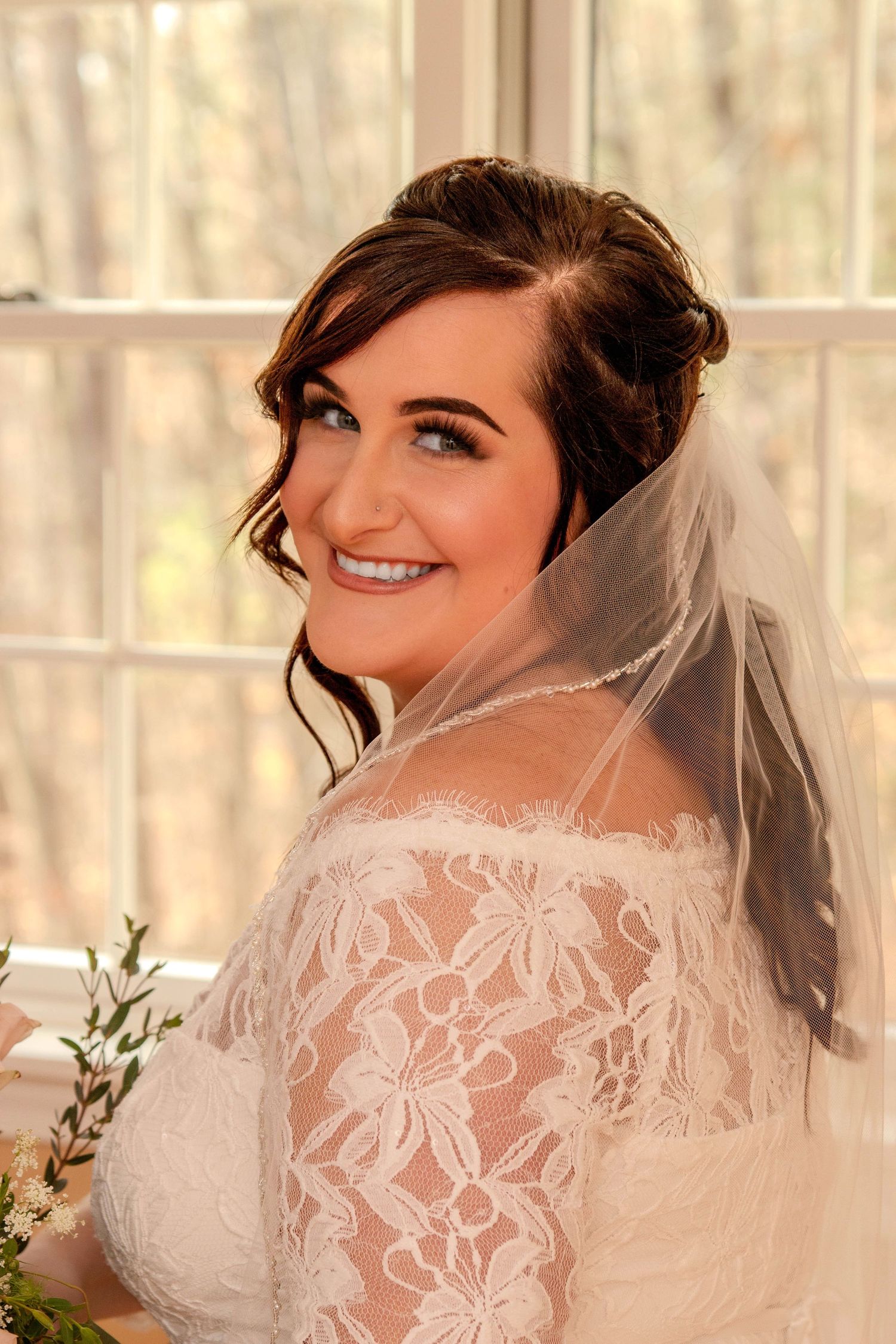 Smiling bride in lace off-shoulder gown and veil poses by sunlit window on wedding day.