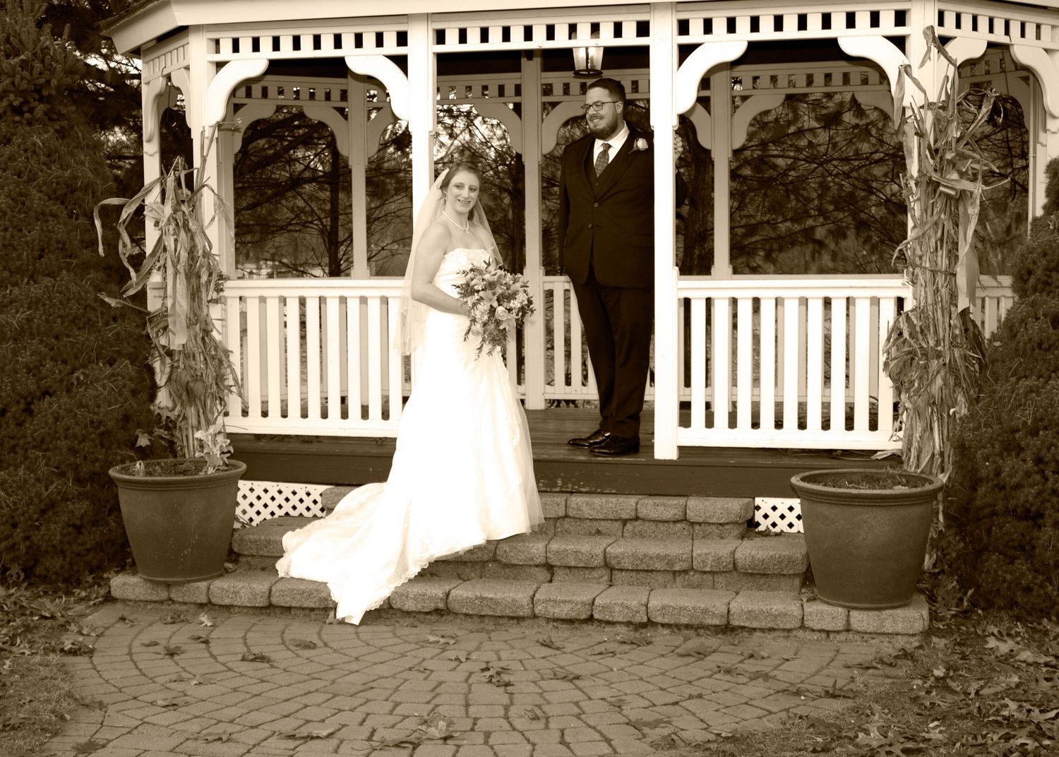 Bride and groom posing at white gazebo steps in sepia-toned vintage wedding portrait.