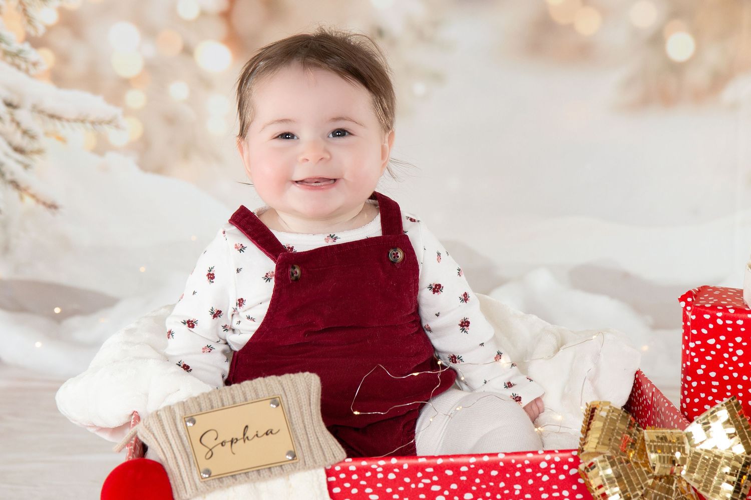 A smiling baby girl named Sophia in red velvet overalls sits beside wrapped Christmas gifts.