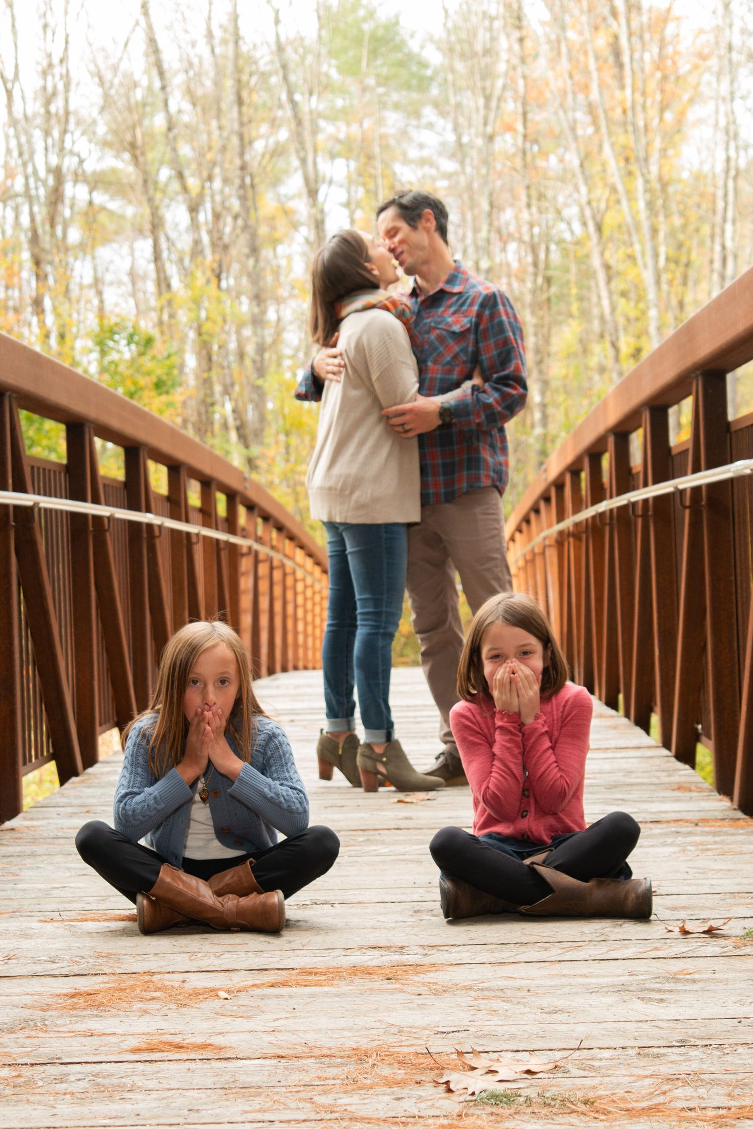 Two girls sit cross-legged on a wooden bridge covering their mouths as their parents kiss behind them in autumn.