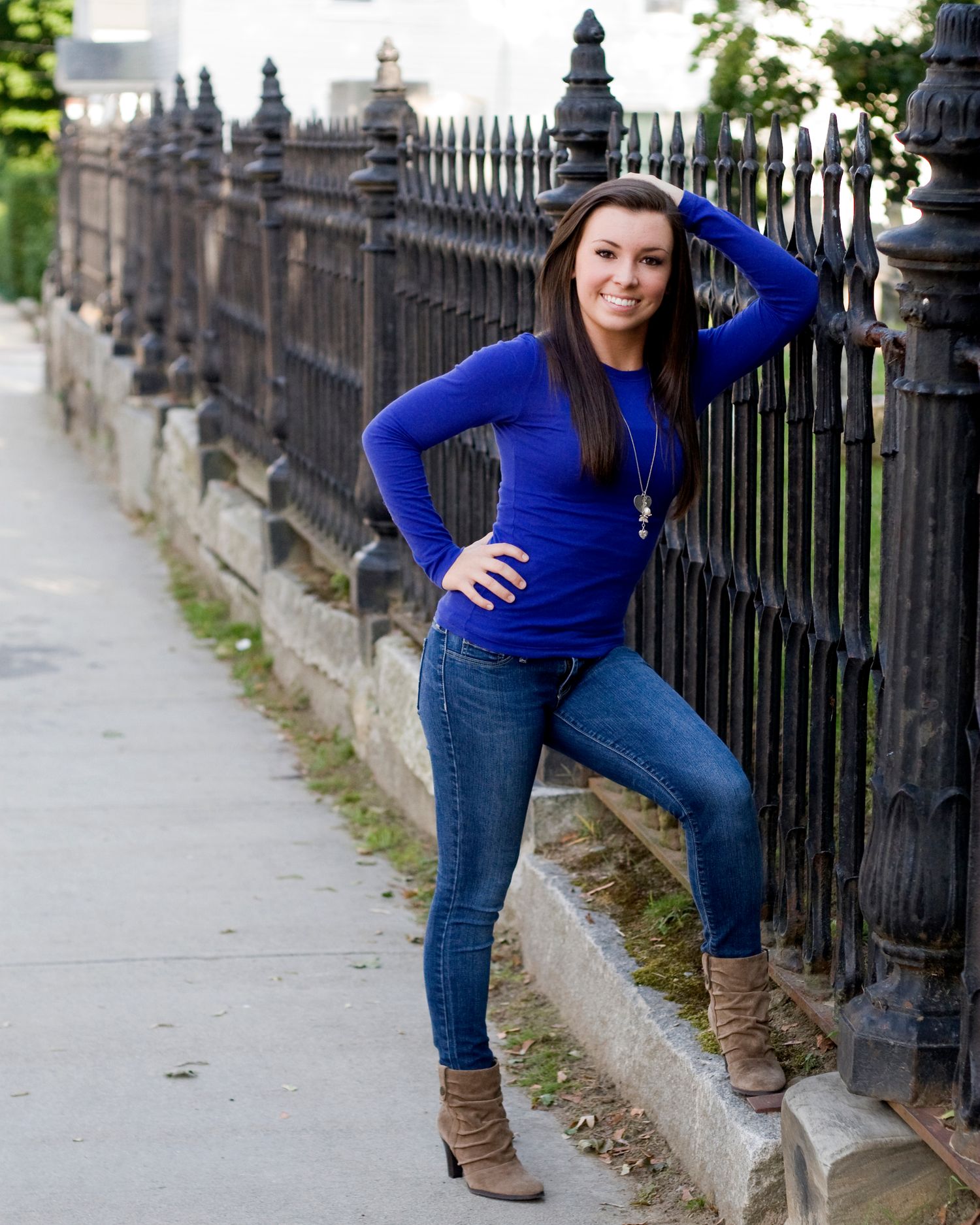Brunette woman in blue top and jeans poses confidently by ornate iron fence outdoors.