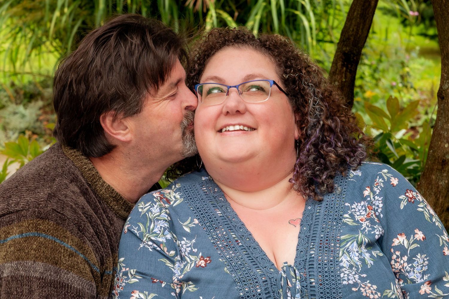 Man kisses smiling woman wearing glasses on the cheek in a lush green garden setting.
