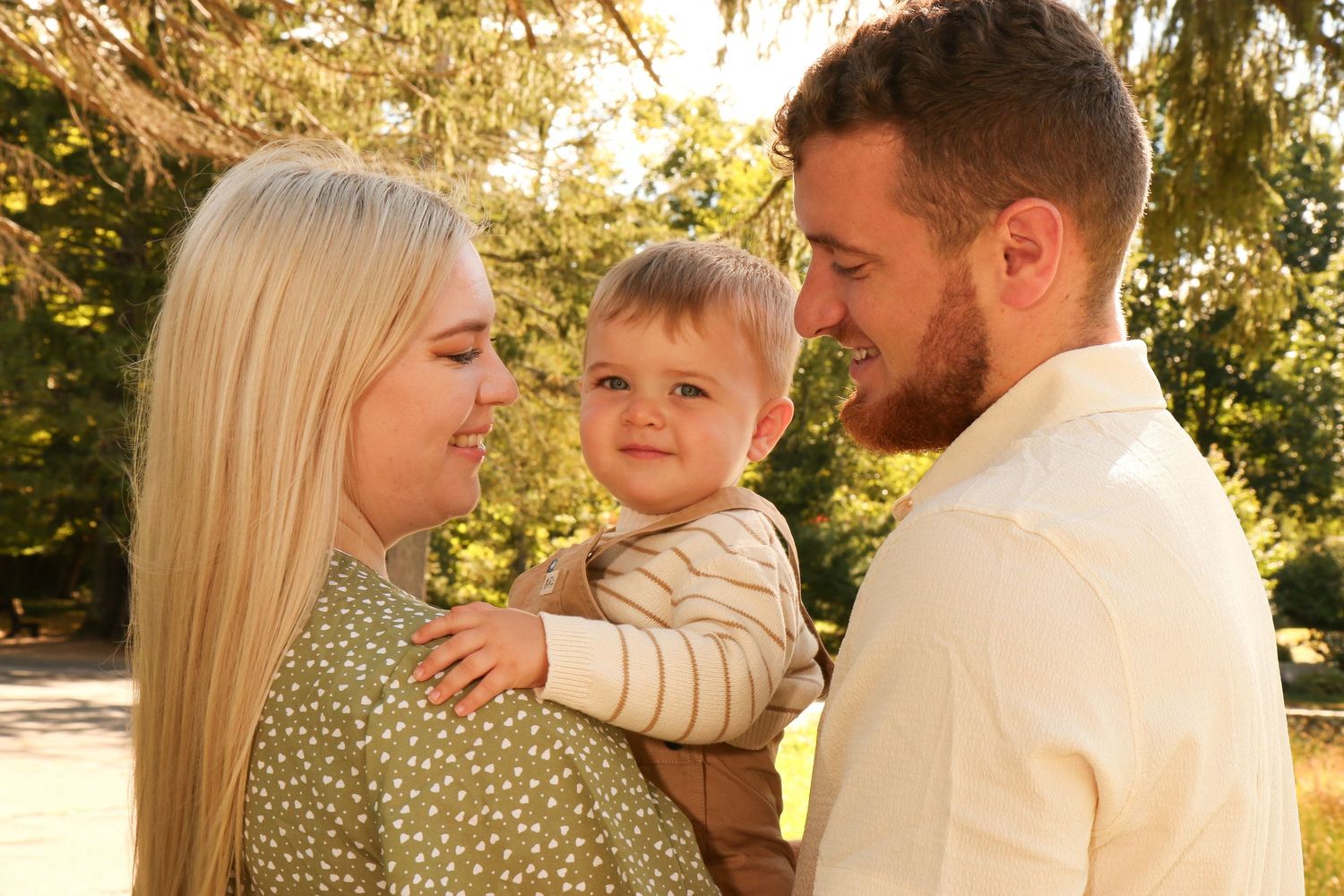 Young family with blonde mother, bearded father, and curious baby boy enjoying sunny outdoor park moment.