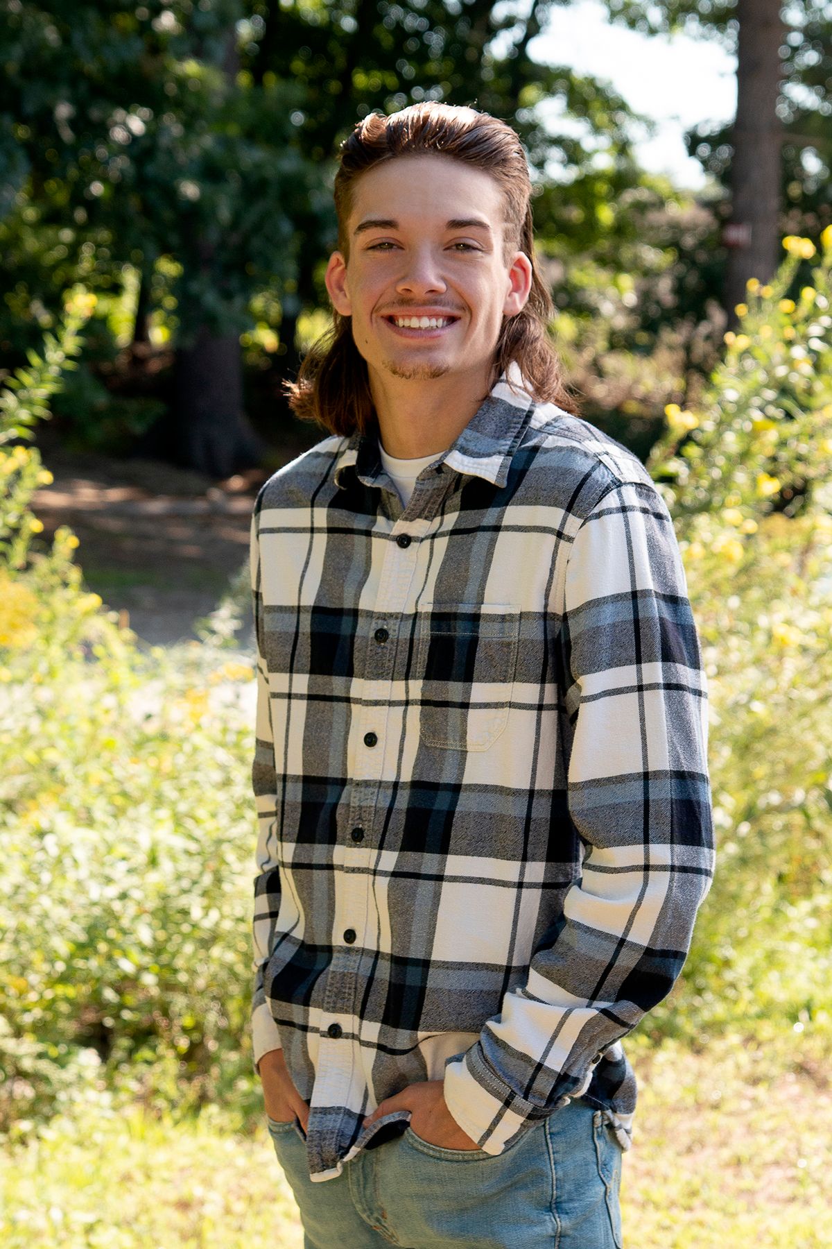 Smiling young man with long hair wearing a plaid flannel shirt standing outdoors in a sunny green garden.