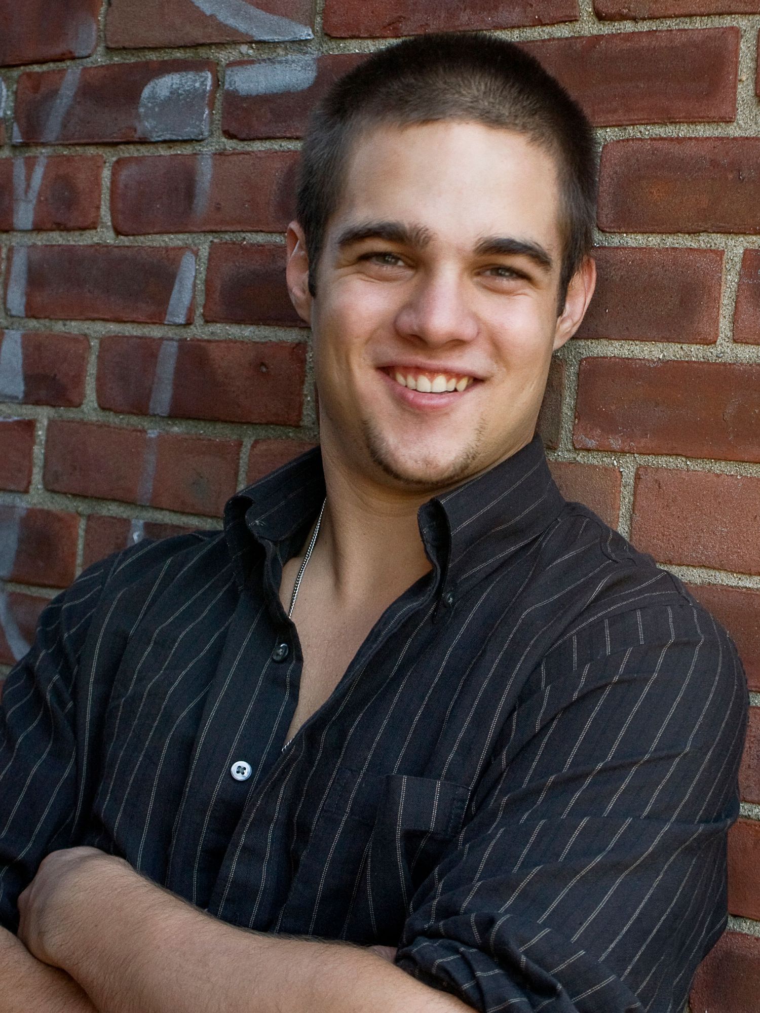 Smiling young man in striped black shirt leans against brick wall with arms crossed.