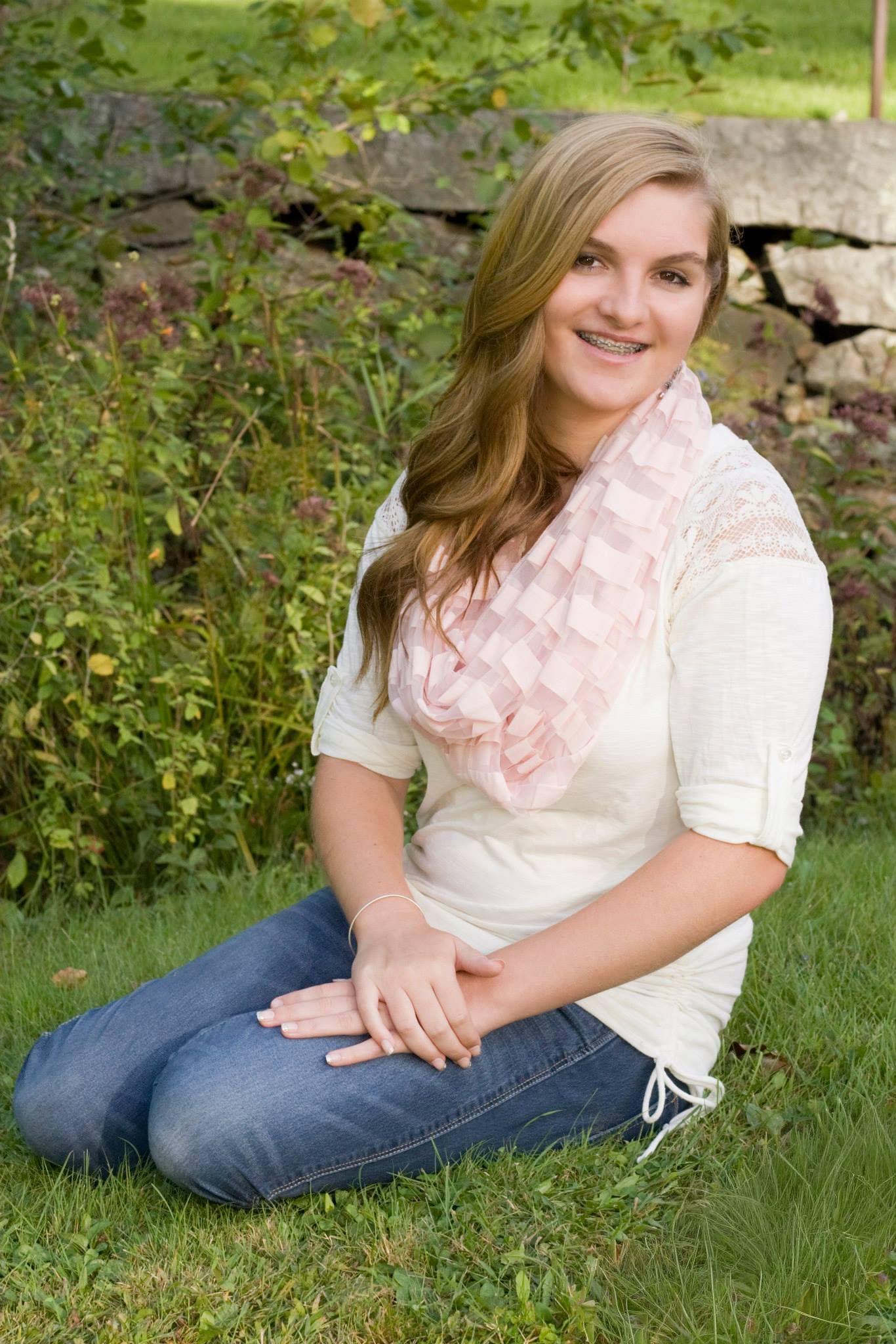 Smiling teenage girl with braces in a cream top and pink scarf sitting on grass in a garden.