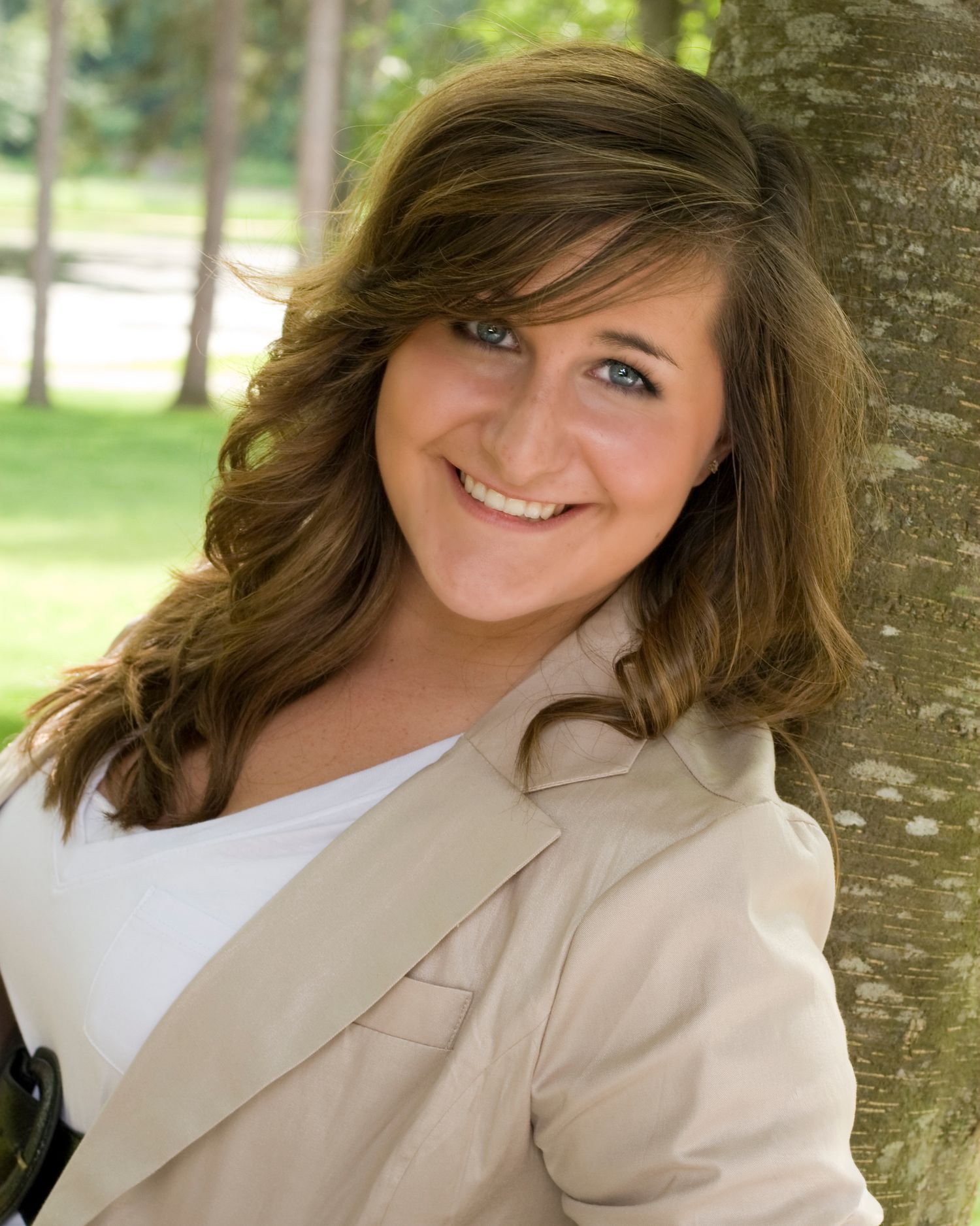 Smiling brunette woman in beige blazer leans against tree trunk in sunny park.