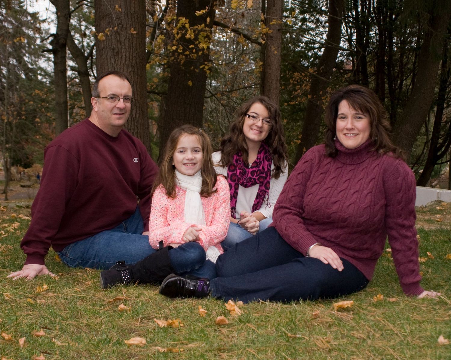 Family of four sits together on grass among autumn trees in outdoor portrait.