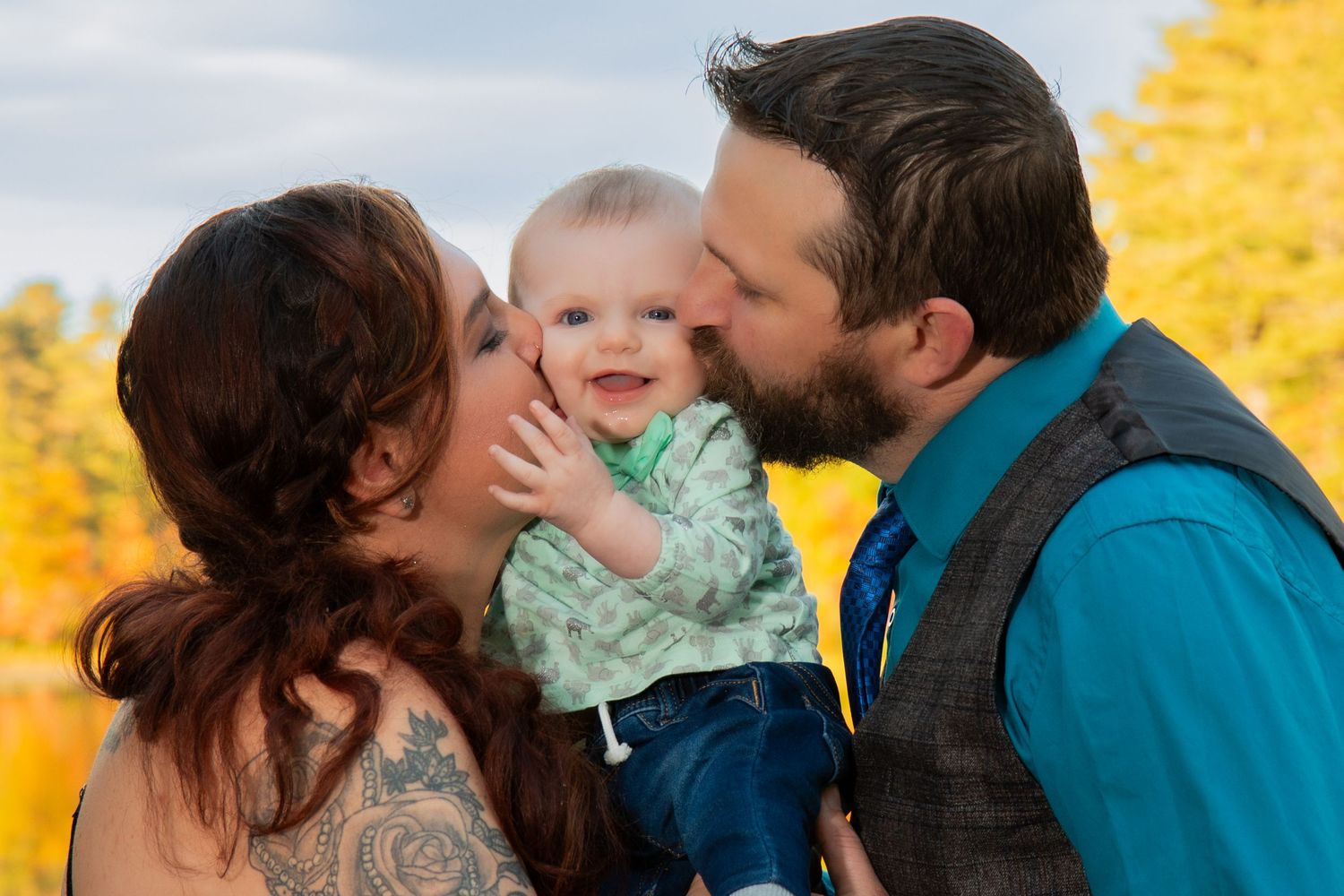 Happy parents kiss their smiling baby boy on the cheeks outdoors with colorful autumn foliage behind them.