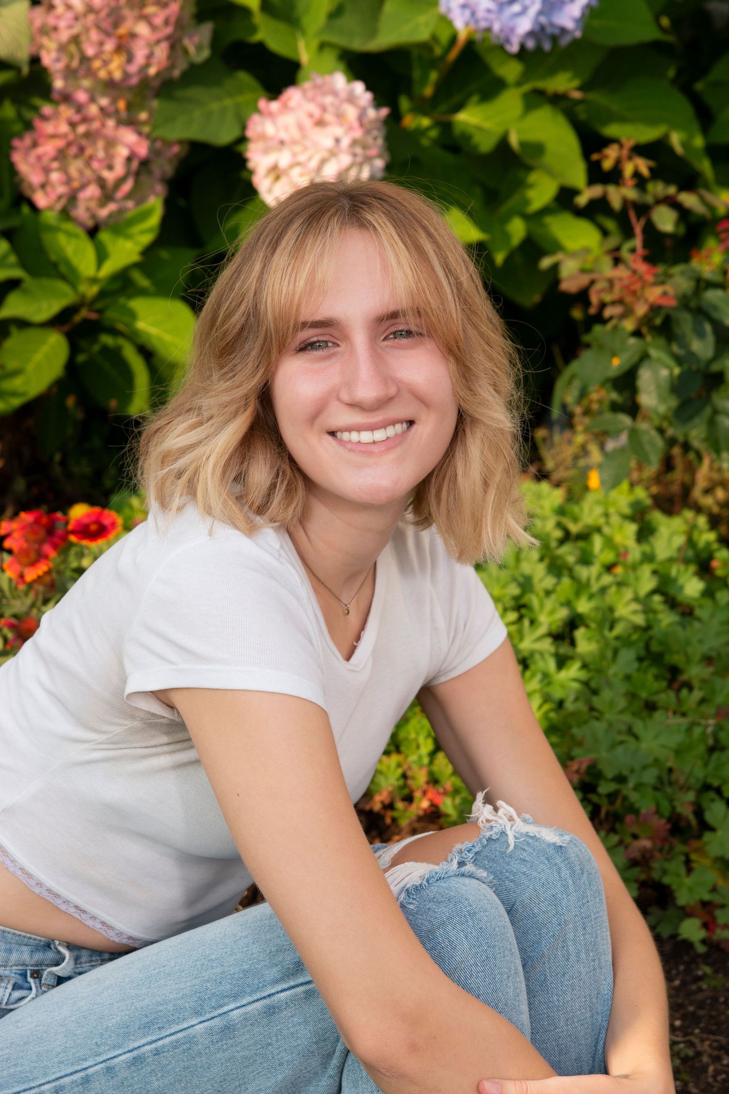 Smiling teenage girl with blonde hair in white t-shirt and jeans posing in garden with hydrangeas.