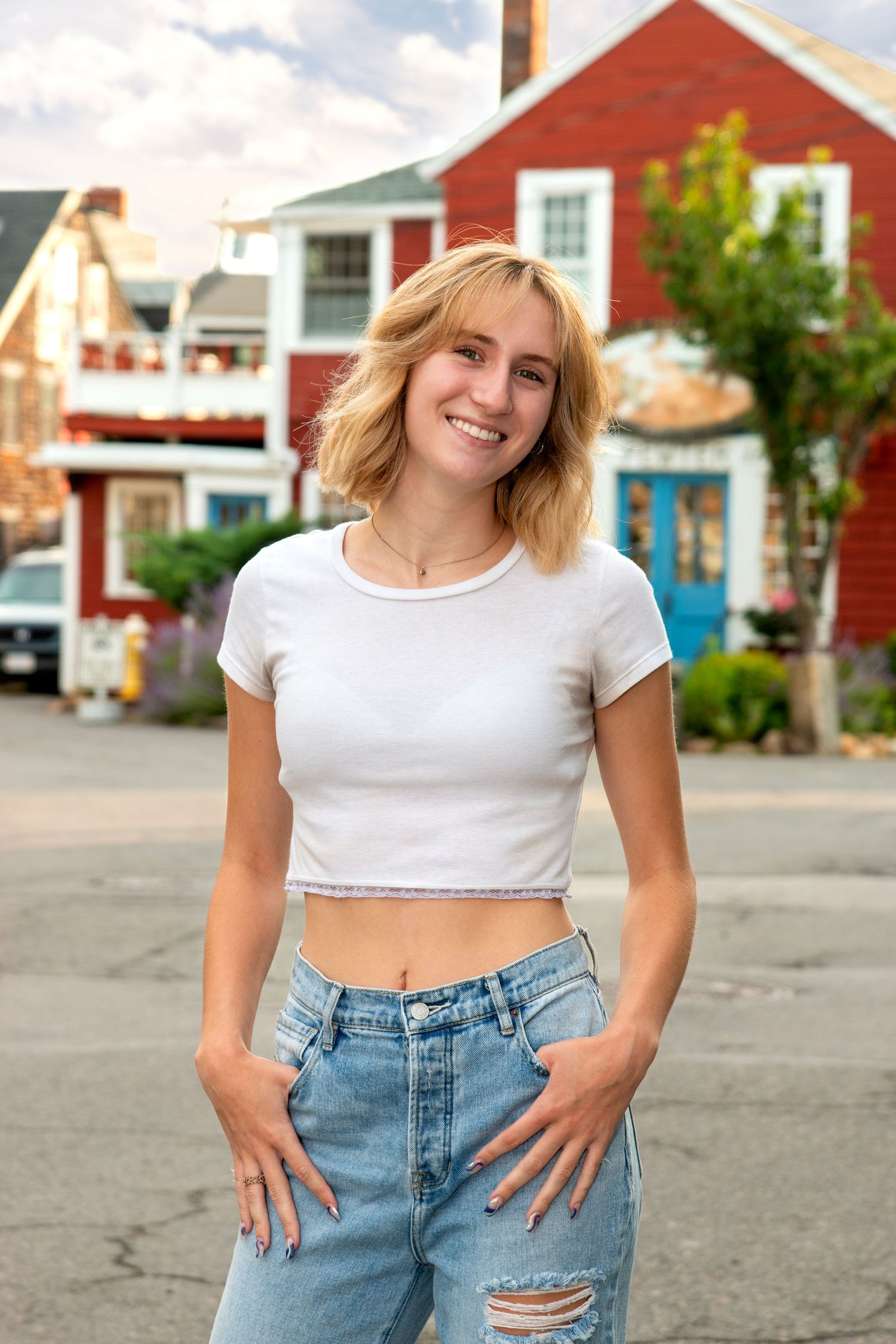 Smiling blonde woman in a white crop top and ripped jeans posing on a street with colorful buildings.