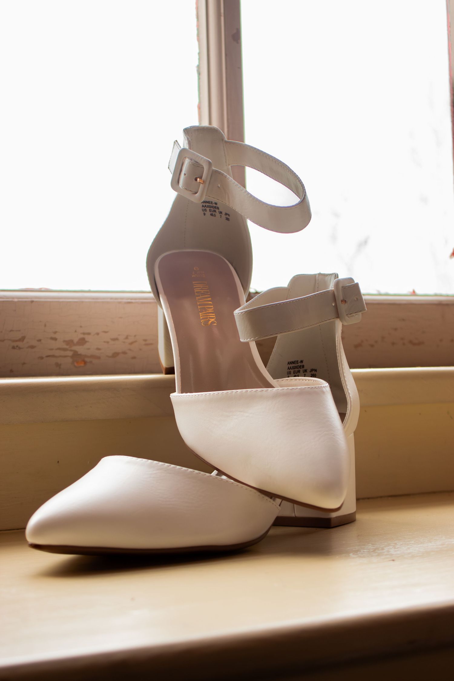 White ankle-strap block heel bridal shoes displayed on wooden windowsill in soft light.