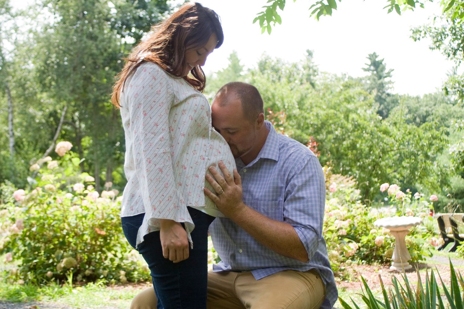 Expectant father kneeling and kissing pregnant partners baby bump in lush garden setting.