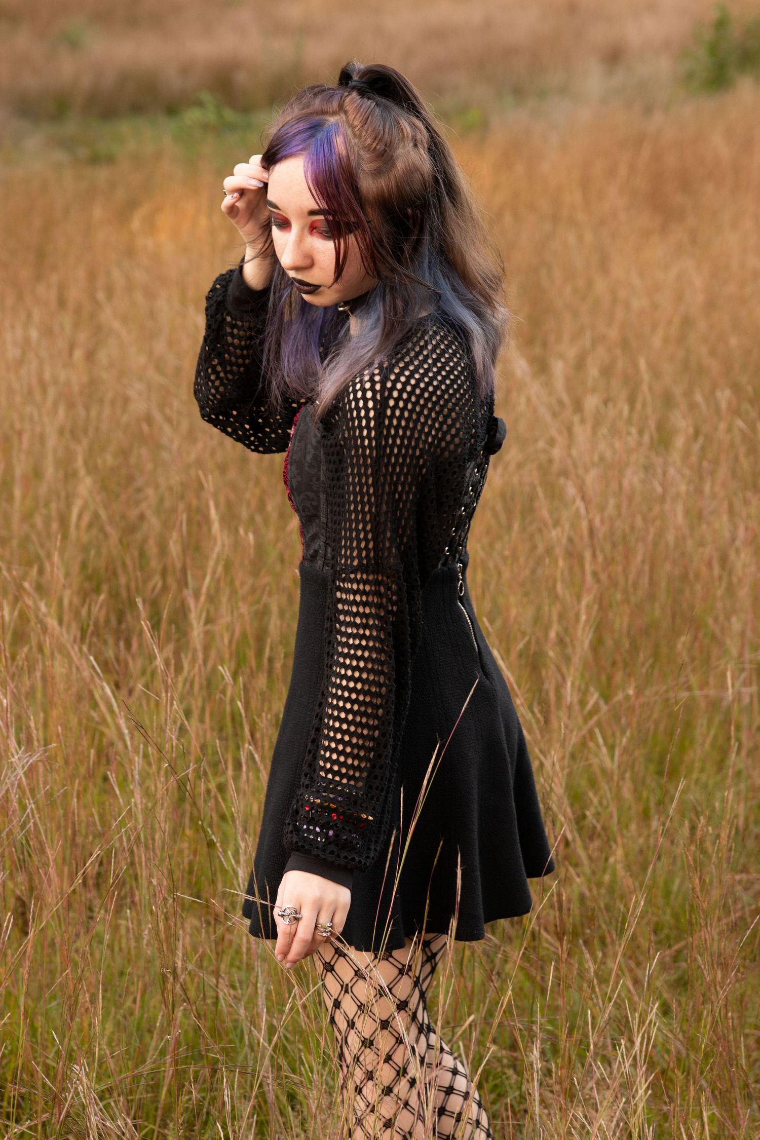 Young woman with purple-tinted hair in black mesh mini dress and fishnet tights posing in golden field.